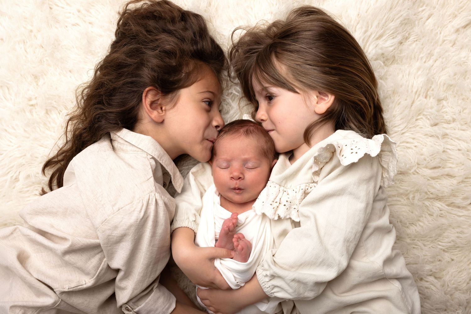 Two young girls in white outfits cuddle a sleeping newborn baby on a soft white fur blanket.