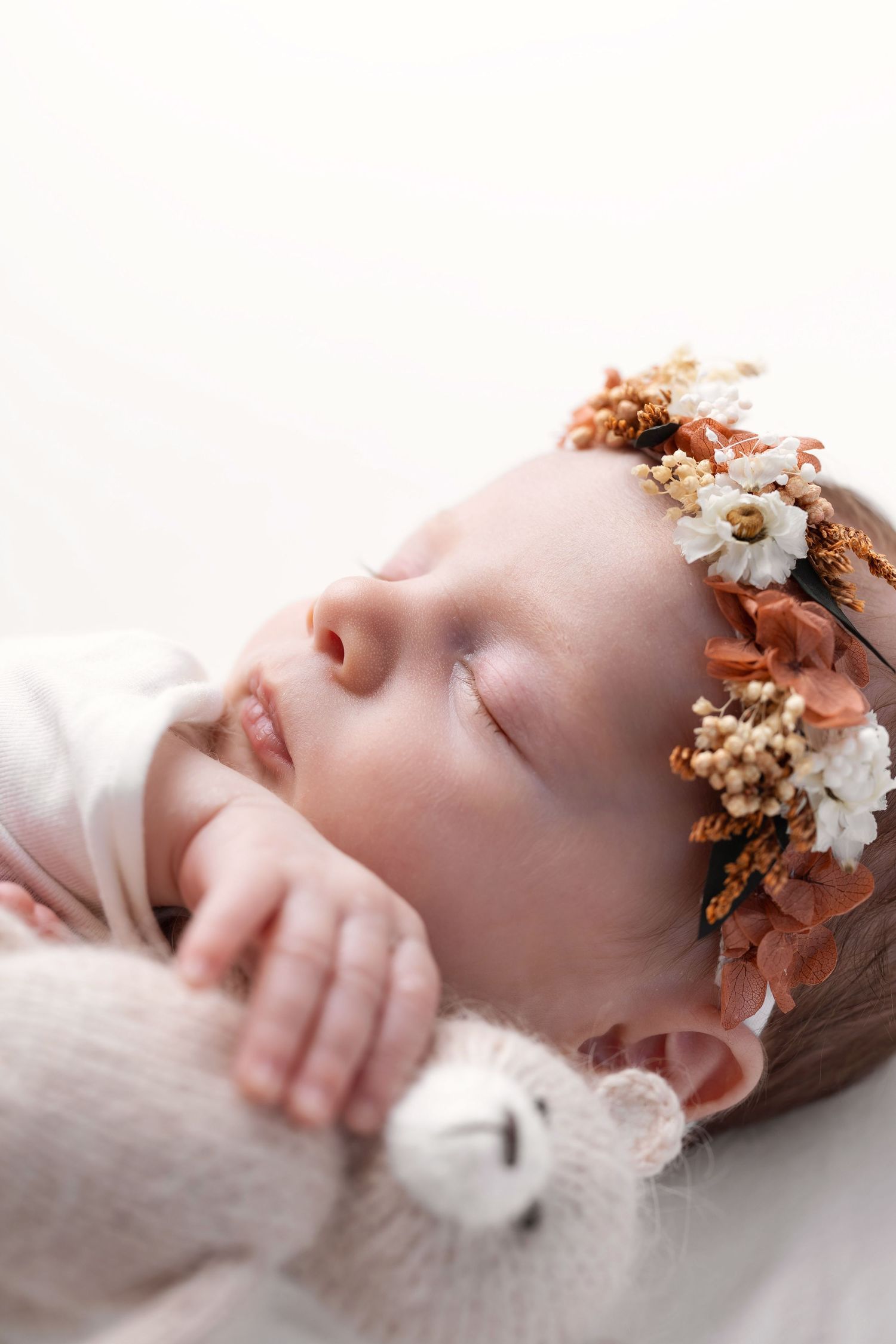 Close-up portrait of sleeping newborn with dried floral crown gently holding a soft white stuffed bear.