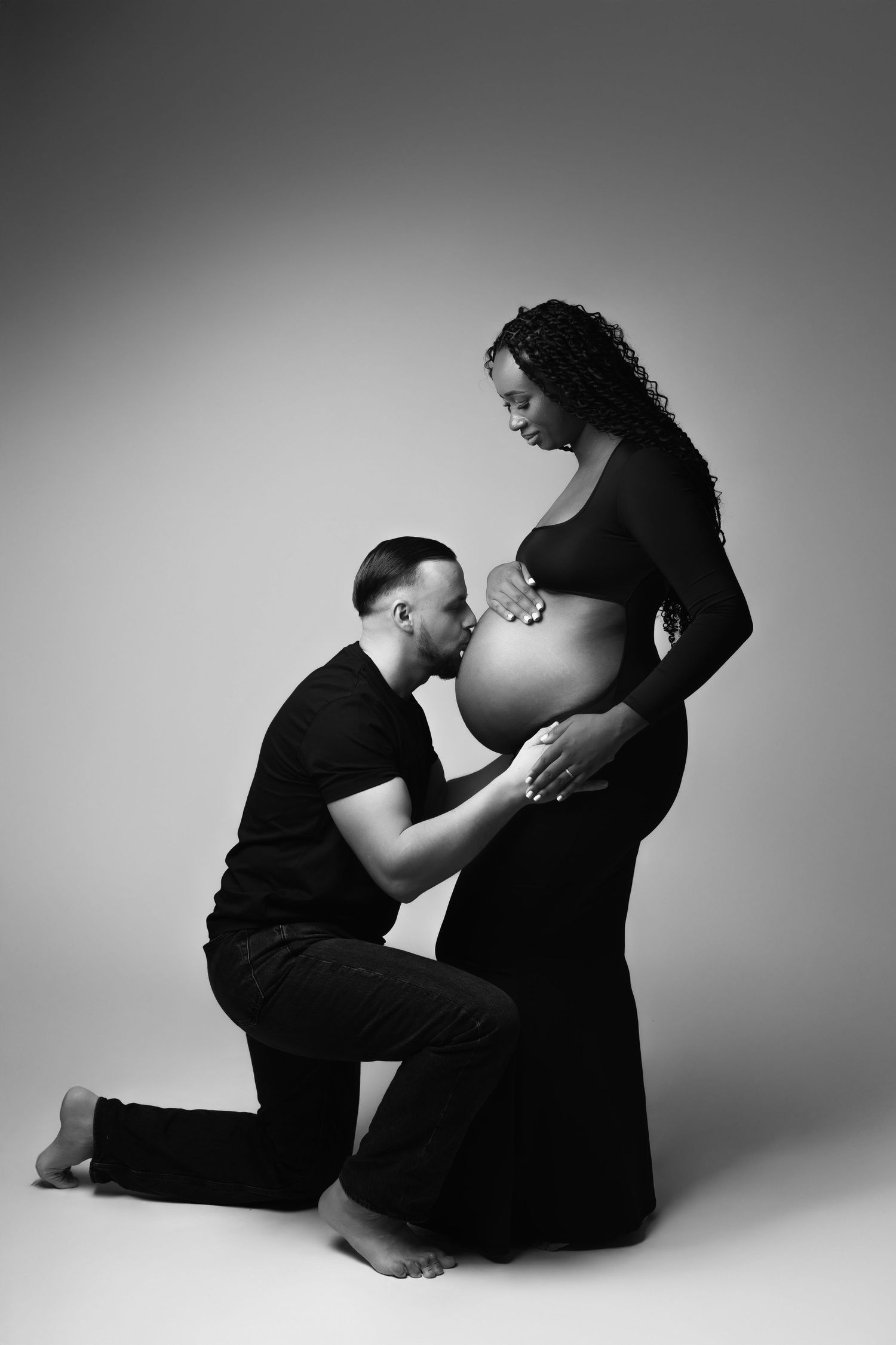 Black and white maternity photo of man kneeling kissing pregnant woman's belly in studio. photographe grossesse chavanoz lyon