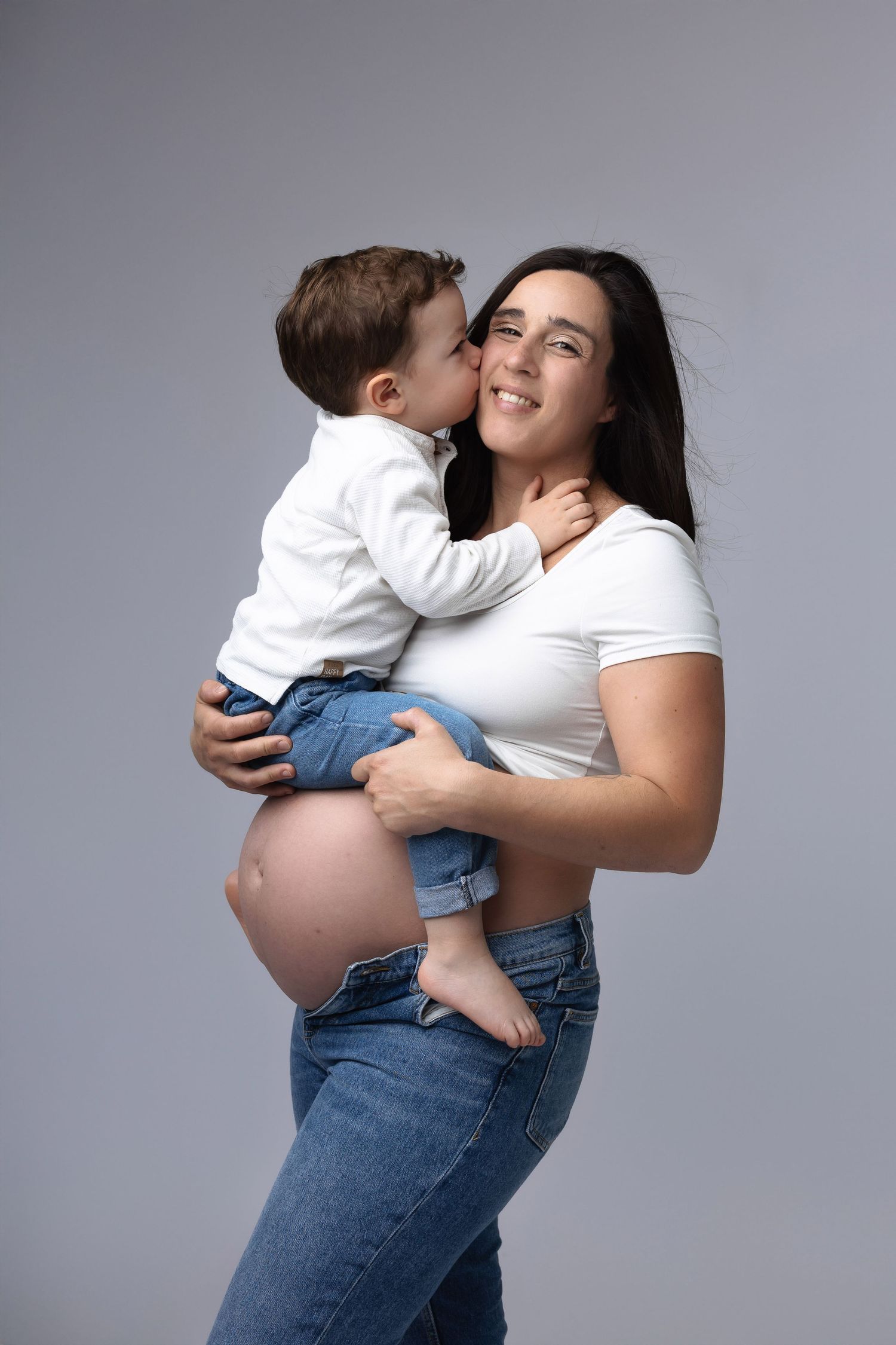 Pregnant woman in white top holding toddler son who kisses her cheek on gray background. photographe grossesse chavanoz lyon