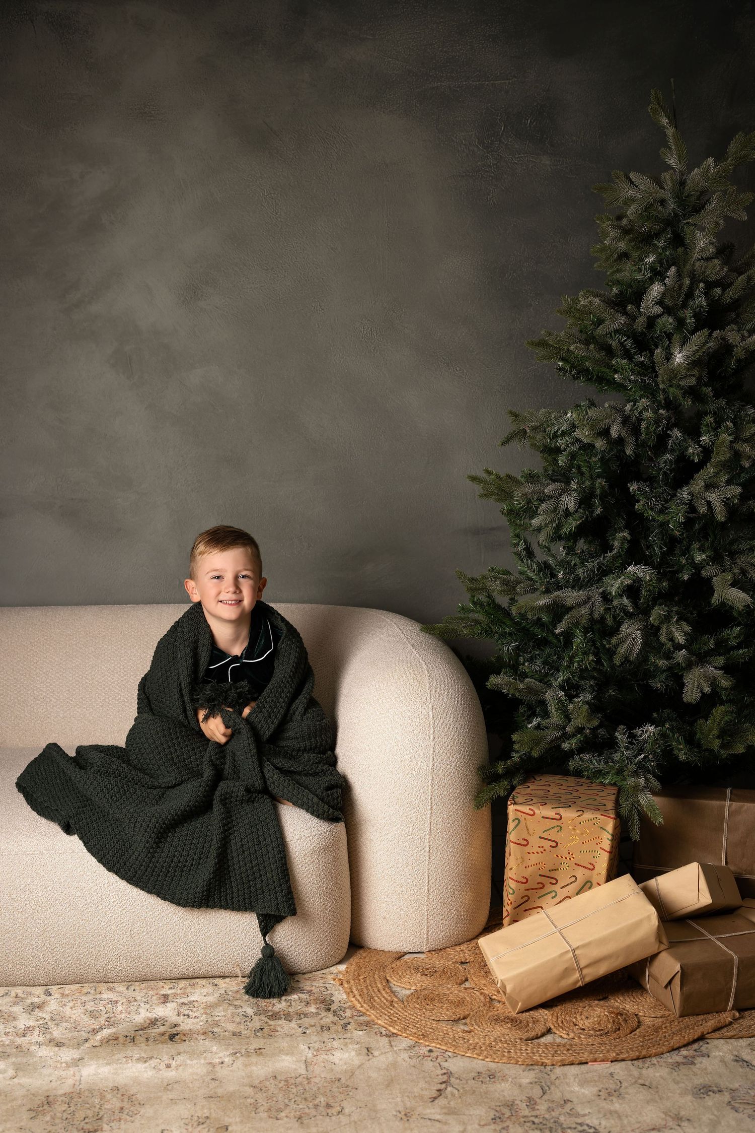 A smiling boy wrapped in a dark knit blanket sits on a beige sofa near a Christmas tree with gifts.