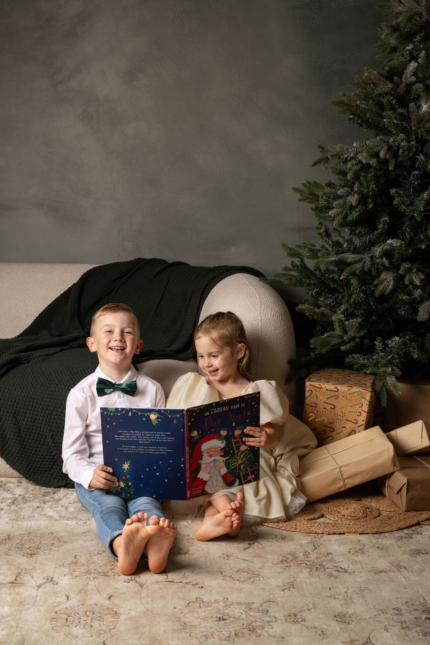 Two happy children sit barefoot by a Christmas tree reading a festive book surrounded by wrapped gifts.