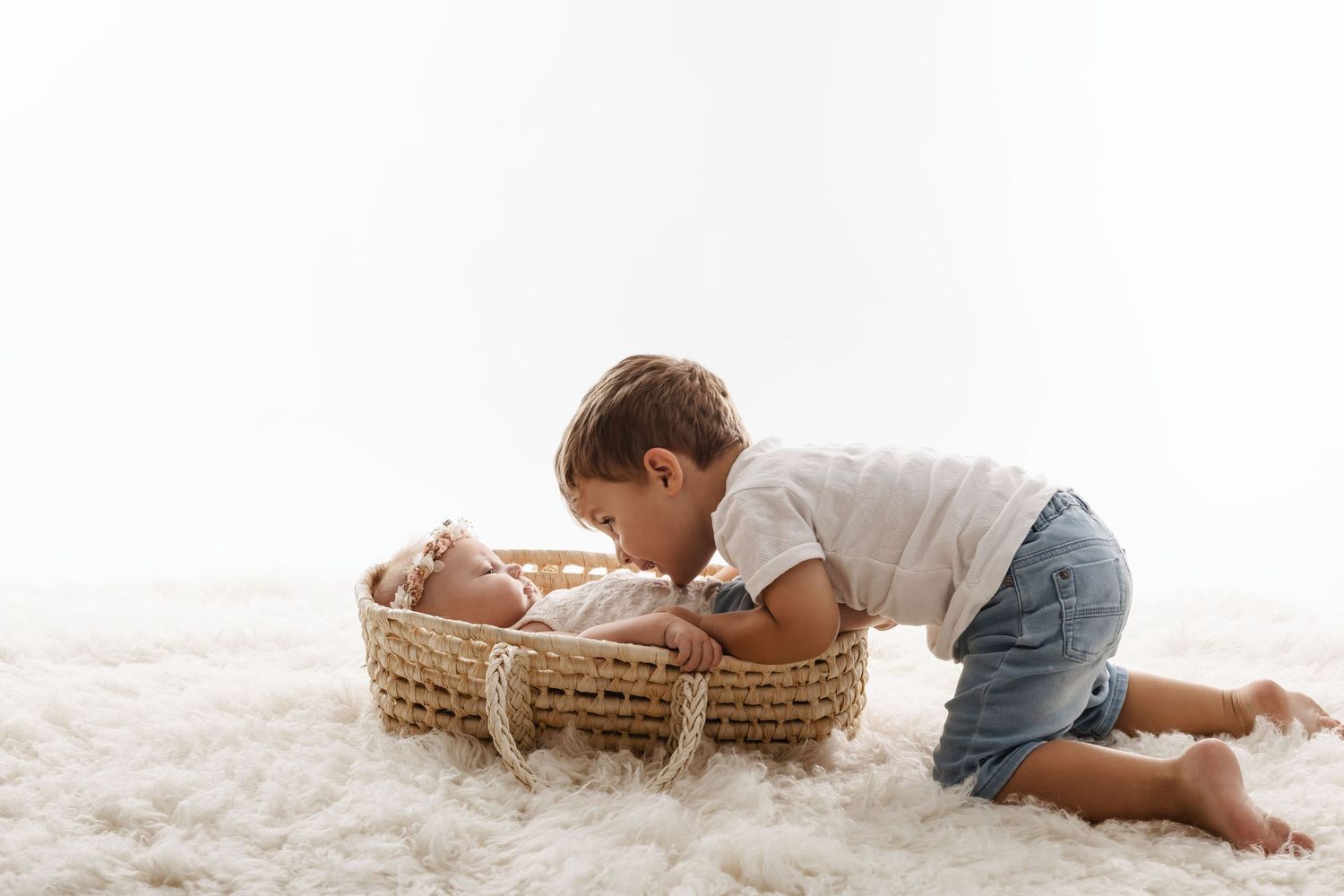 A young toddler boy kisses his newborn sibling resting in a wicker basket on a soft white fluffy rug.