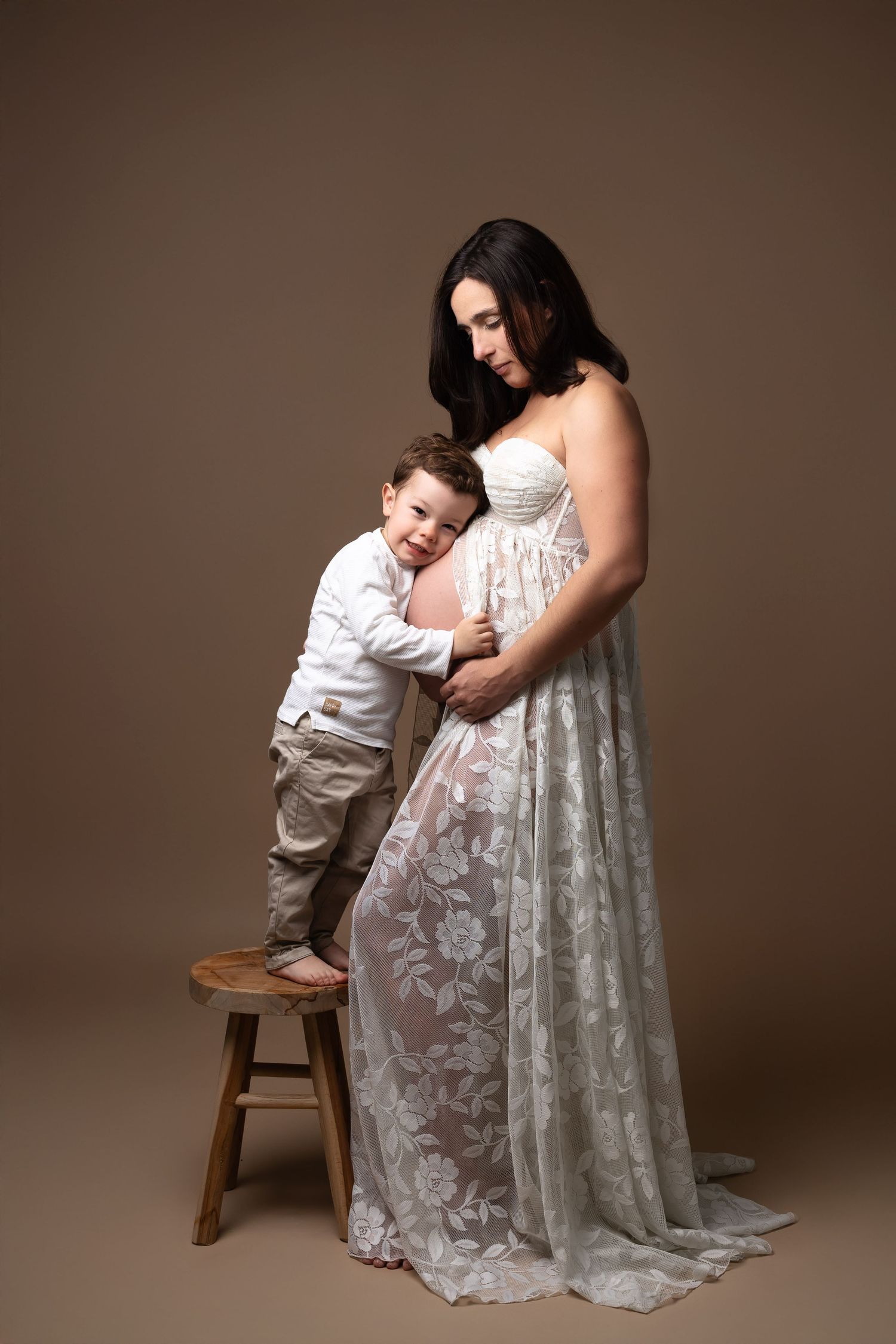 Pregnant woman in white lace gown with toddler son standing on stool hugging her belly. photographe grossesse chavanoz lyon