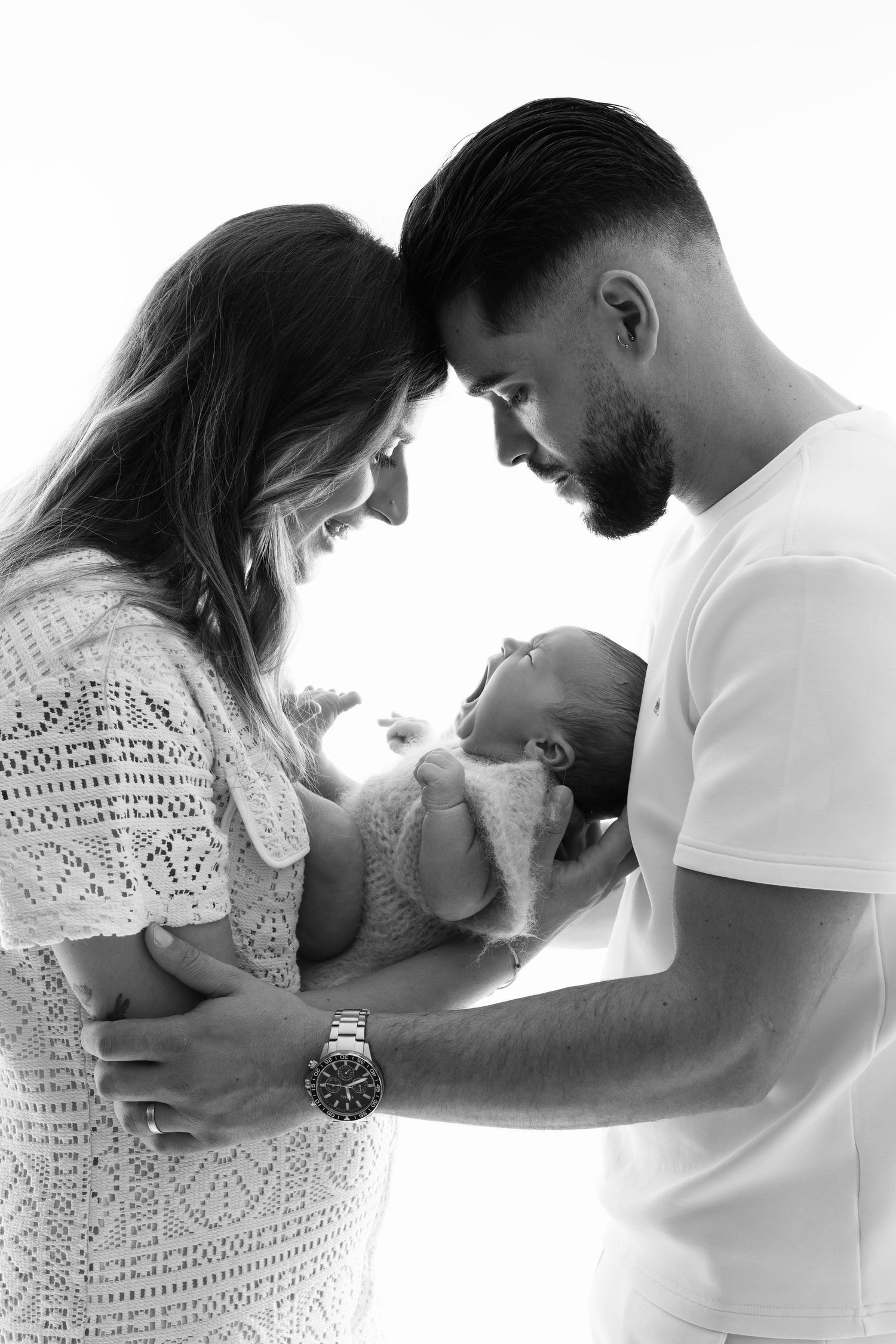 Black and white photo of parents smiling at their newborn baby, touching foreheads together in a tender family moment.