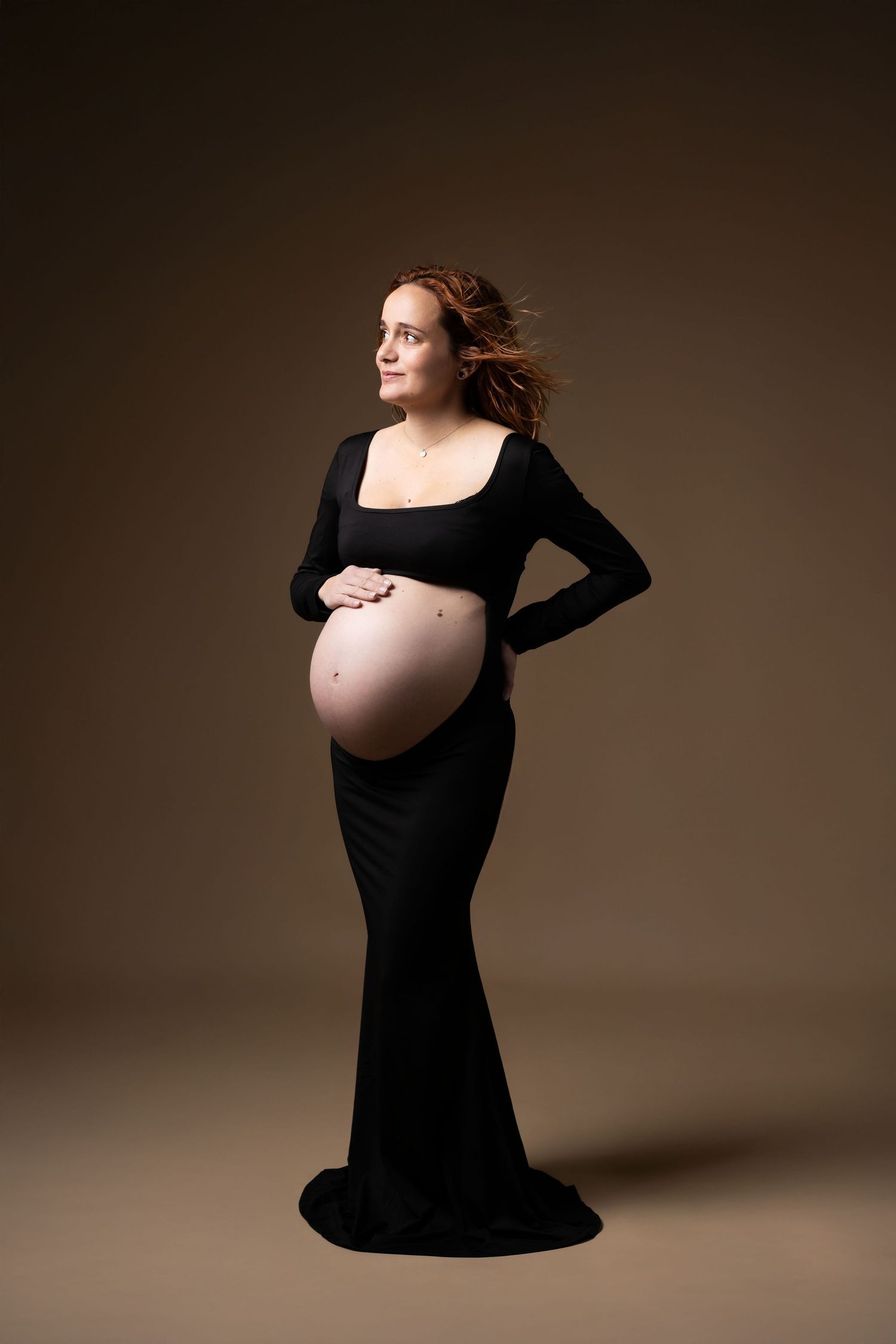 Pregnant woman in black long-sleeve gown with exposed belly, posing against brown background.