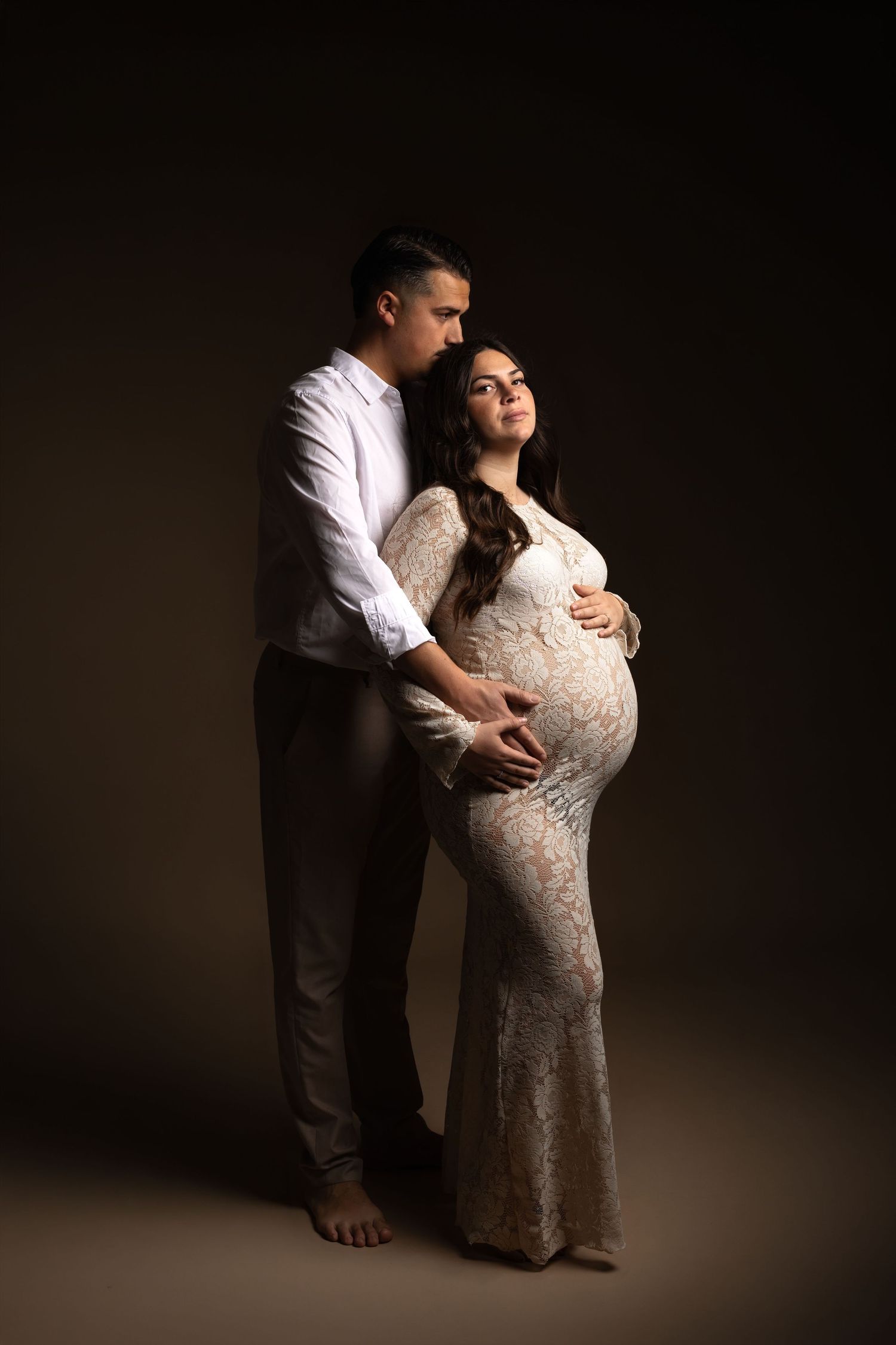 A couple poses for a maternity photo, the woman in a white lace gown cradling her baby bump, her partner embracing her from behind.