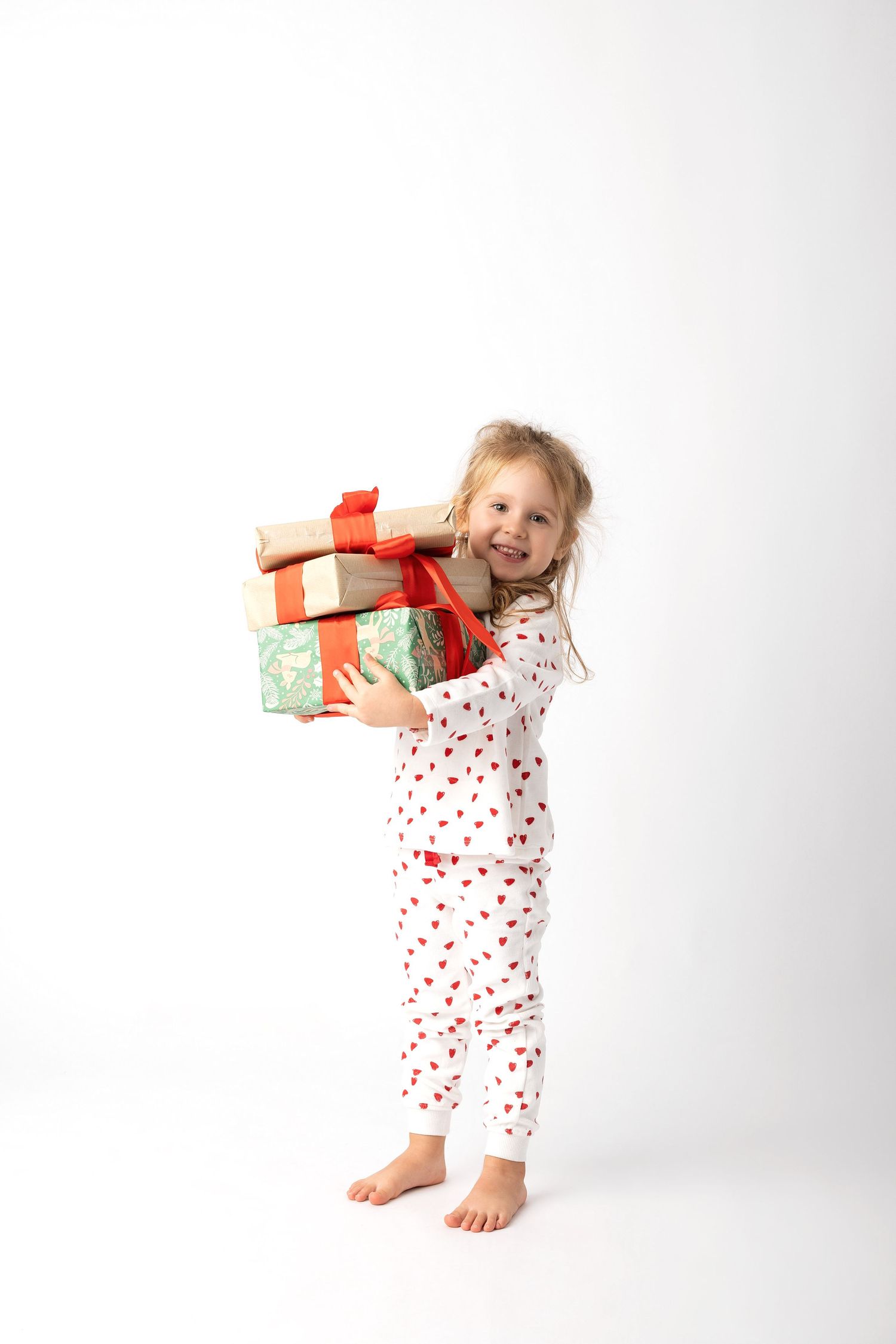 Young girl in heart-print pajamas holding stacked Christmas gifts with red ribbons, smiling against white background.