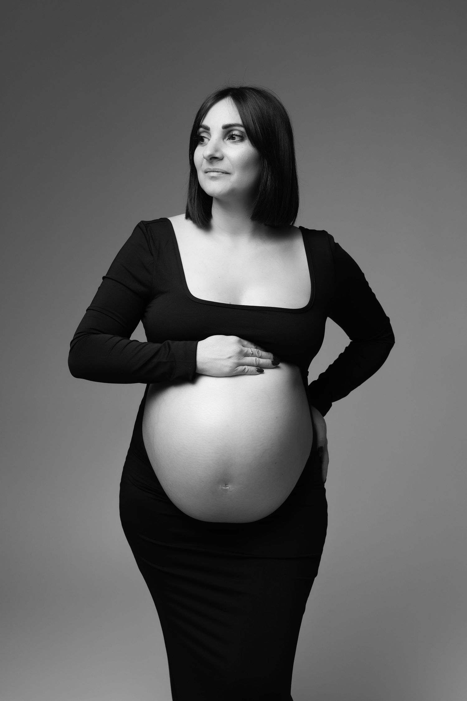 Black and white studio portrait of pregnant brunette woman in black dress cradling her belly. photographe grossesse chavanoz lyon