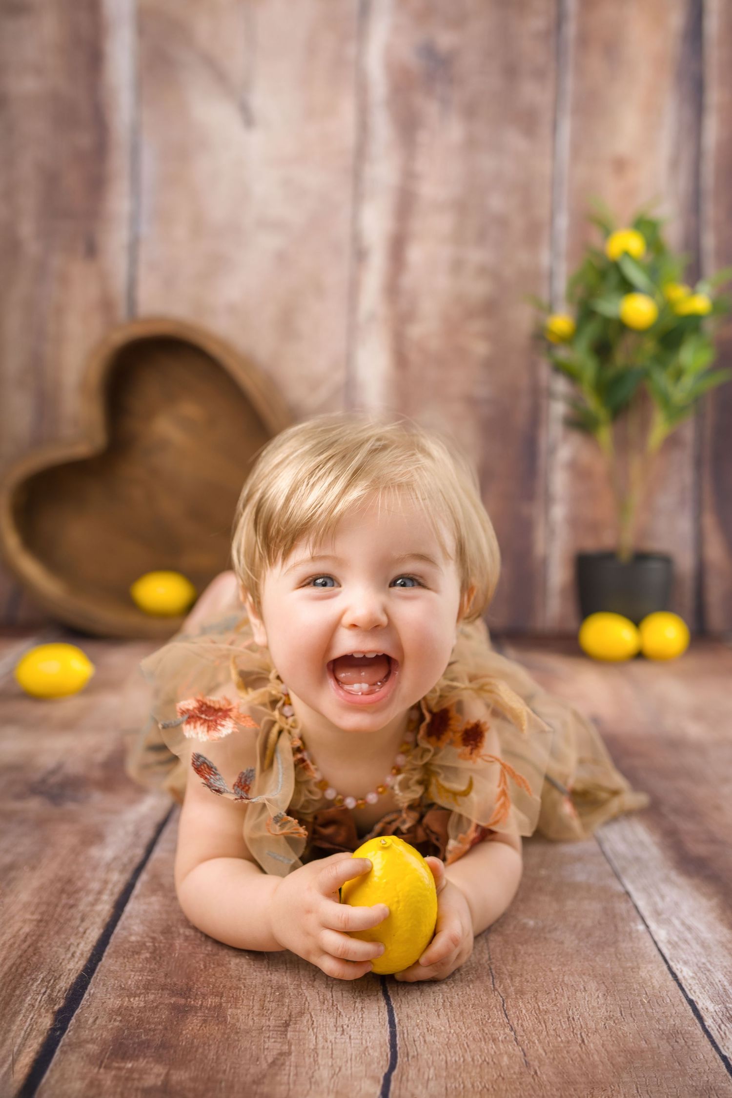 A cheerful baby girl in a brown dress lies on a rustic wood floor holding a yellow lemon.