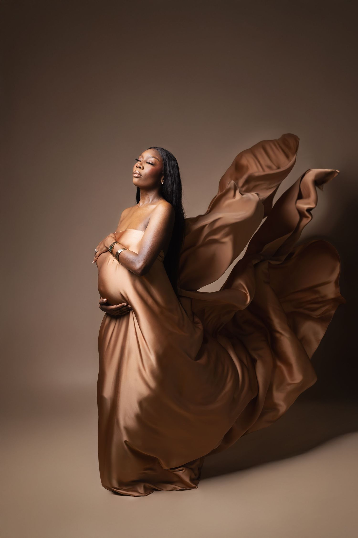 Pregnant Black woman with flowing gold silk fabric billowing in studio.