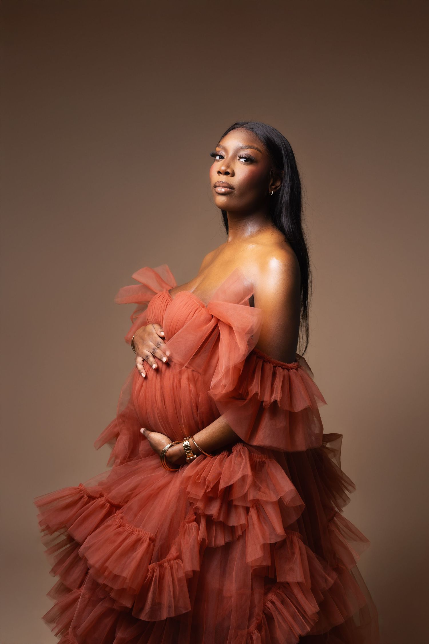 Pregnant Black woman in strapless rust tulle gown posing in studio.