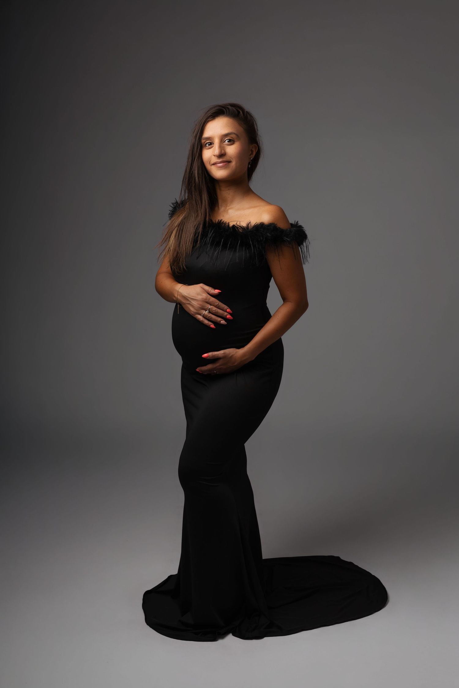 Pregnant woman in elegant black off-shoulder gown with feather trim, cradling baby bump in studio portrait.