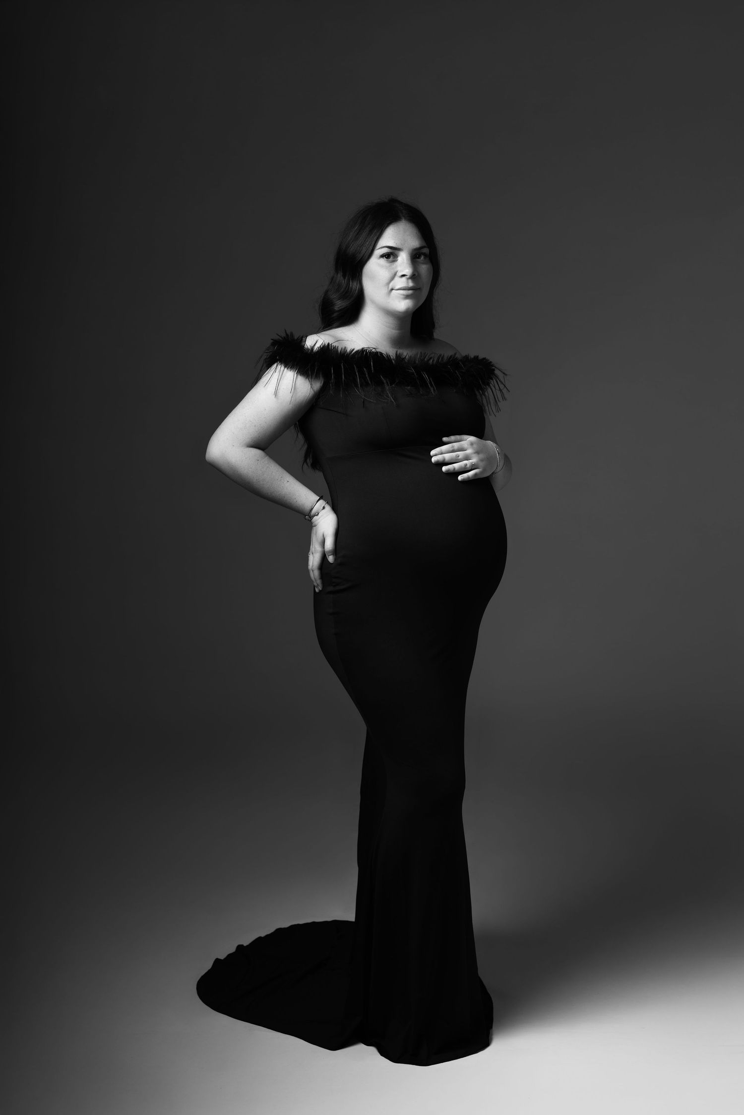 Pregnant woman in elegant black off-shoulder gown with feather trim, posing in professional maternity studio portrait.