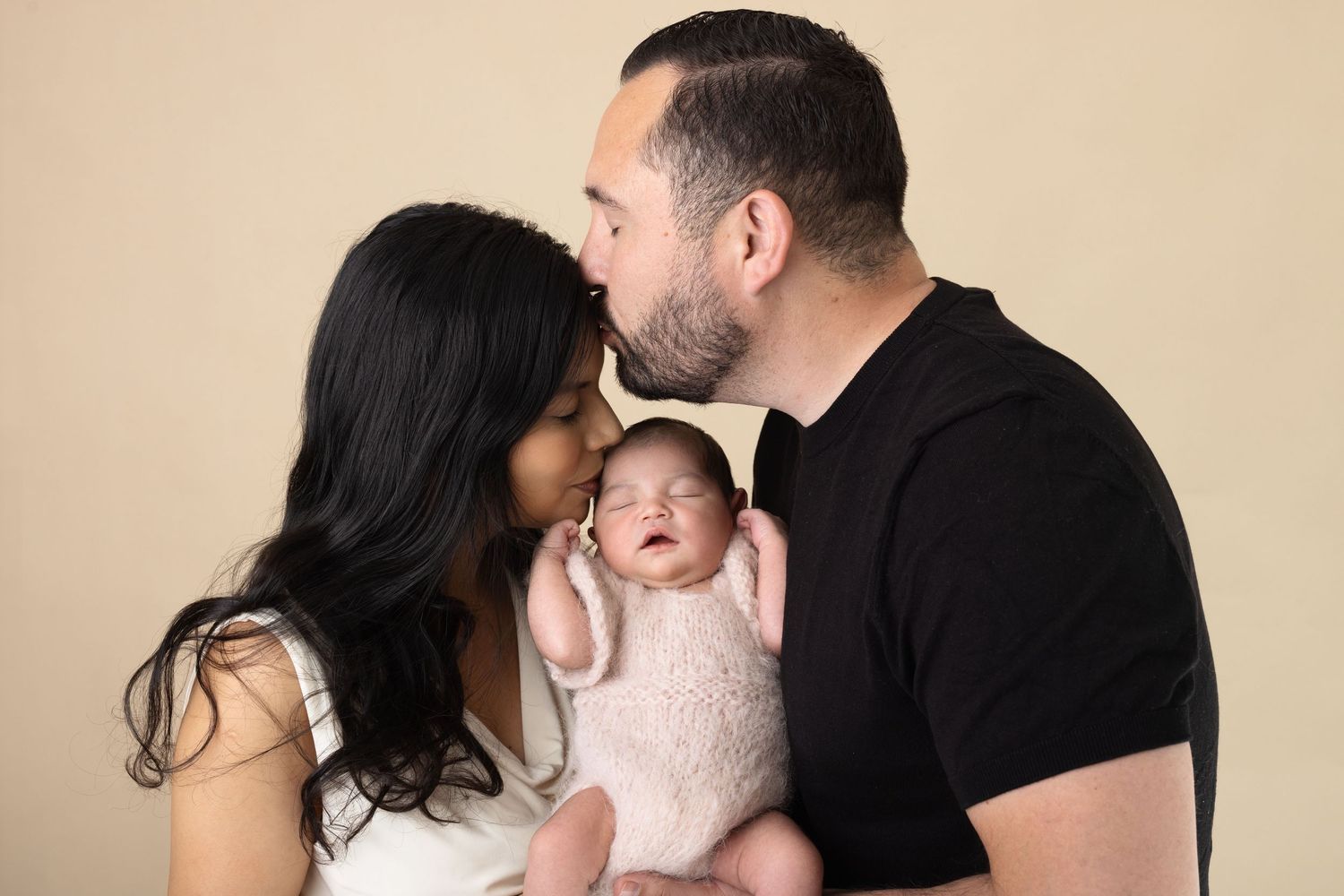 Parents kissing their sleeping newborn baby dressed in pink knit outfit against a beige studio background.