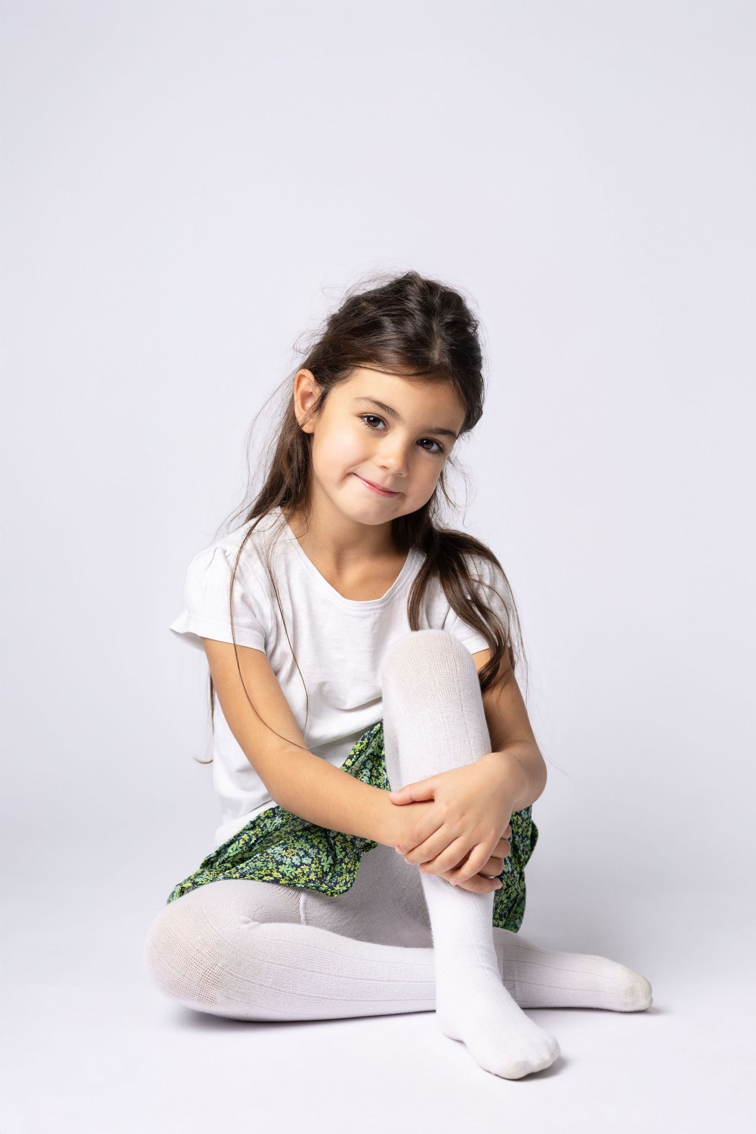 Cute dark-haired girl in white t-shirt and green skirt sits cross-legged wearing white tights on white background.