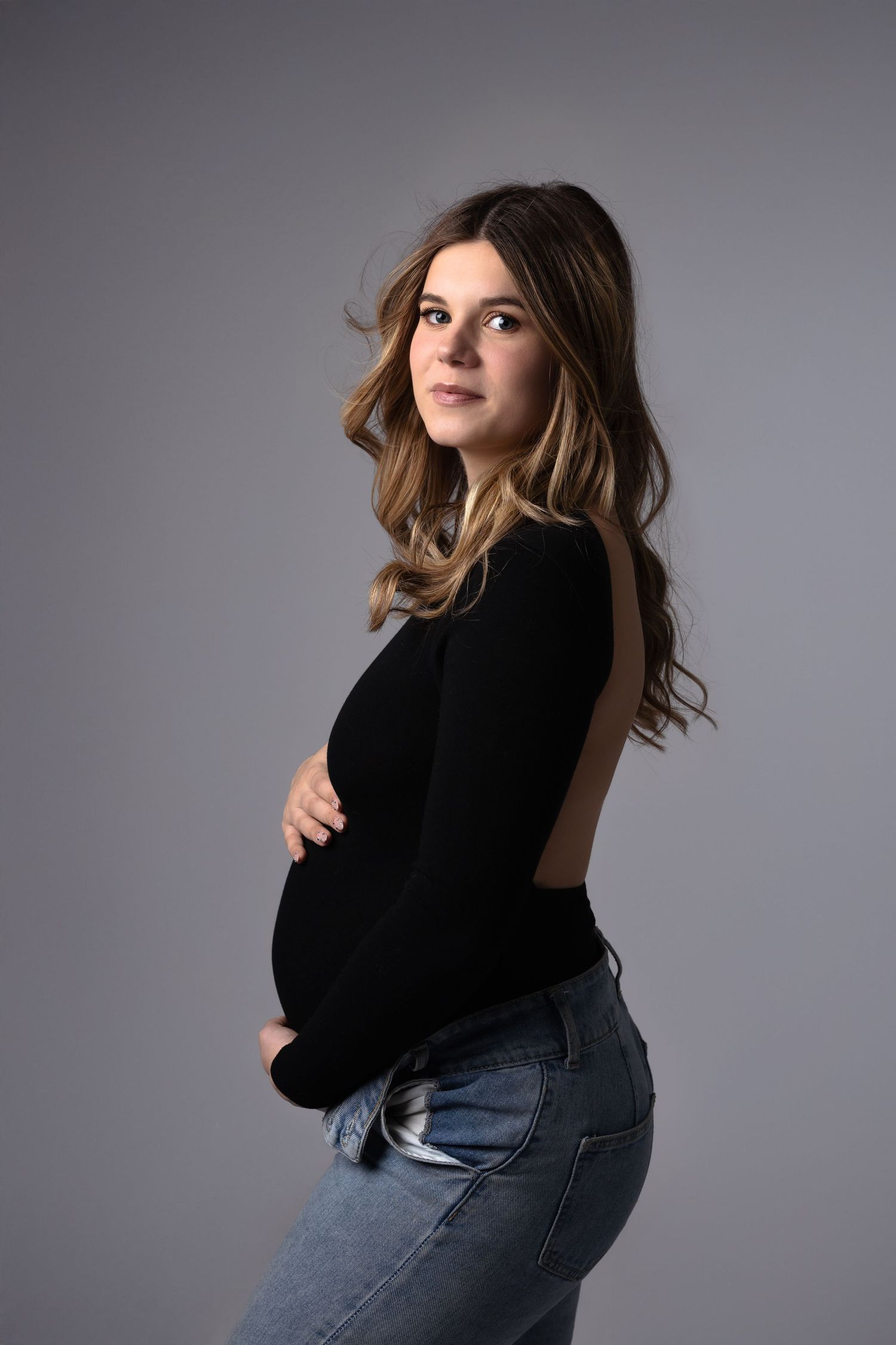 Pregnant woman in black top and jeans posing sideways, cradling baby bump in studio with gray background.