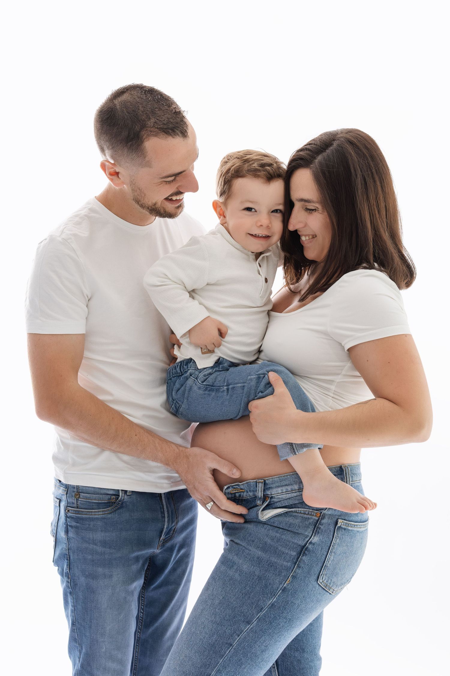 Happy family with mother, father, and baby boy all wearing white shirts and jeans posing together. photographe grossesse chavanoz lyon