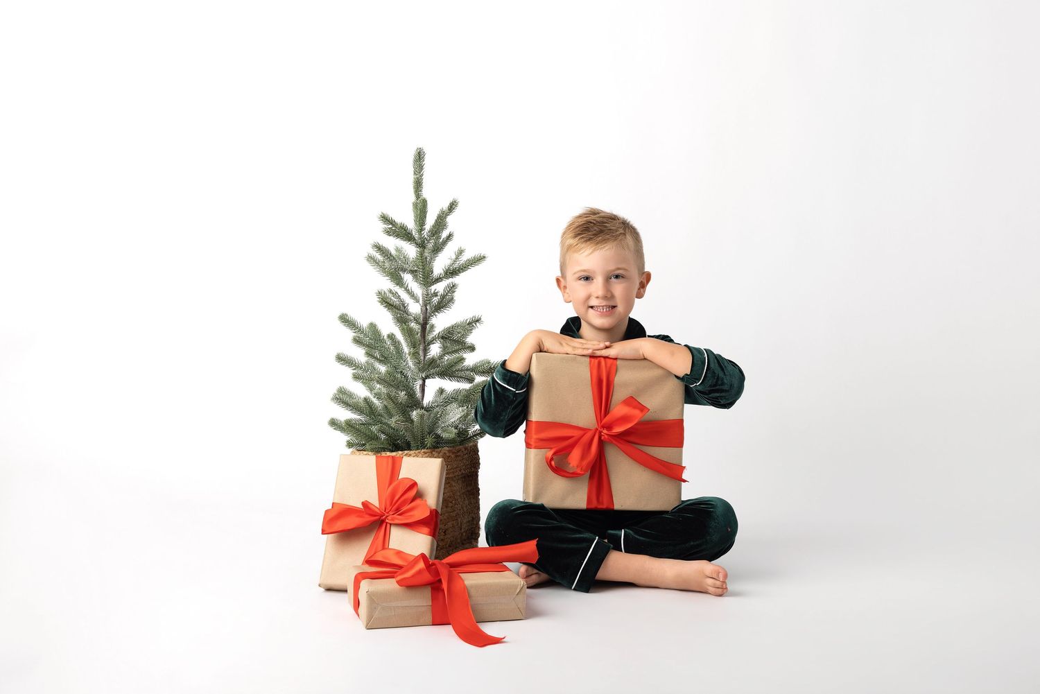 Smiling boy sitting cross-legged holding a gift box with red ribbon near a small Christmas tree and wrapped presents.