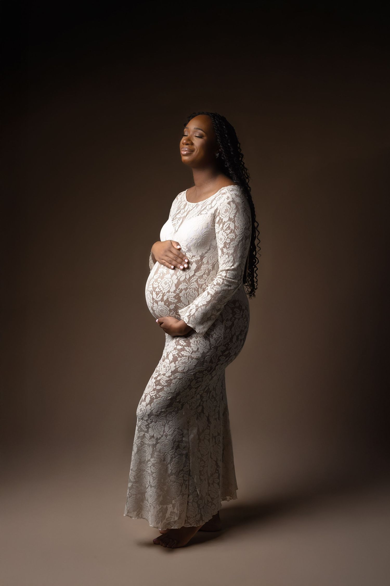 Pregnant Black woman in white lace gown cradling baby bump, smiling in studio portrait. photographe grossesse chavanoz lyon