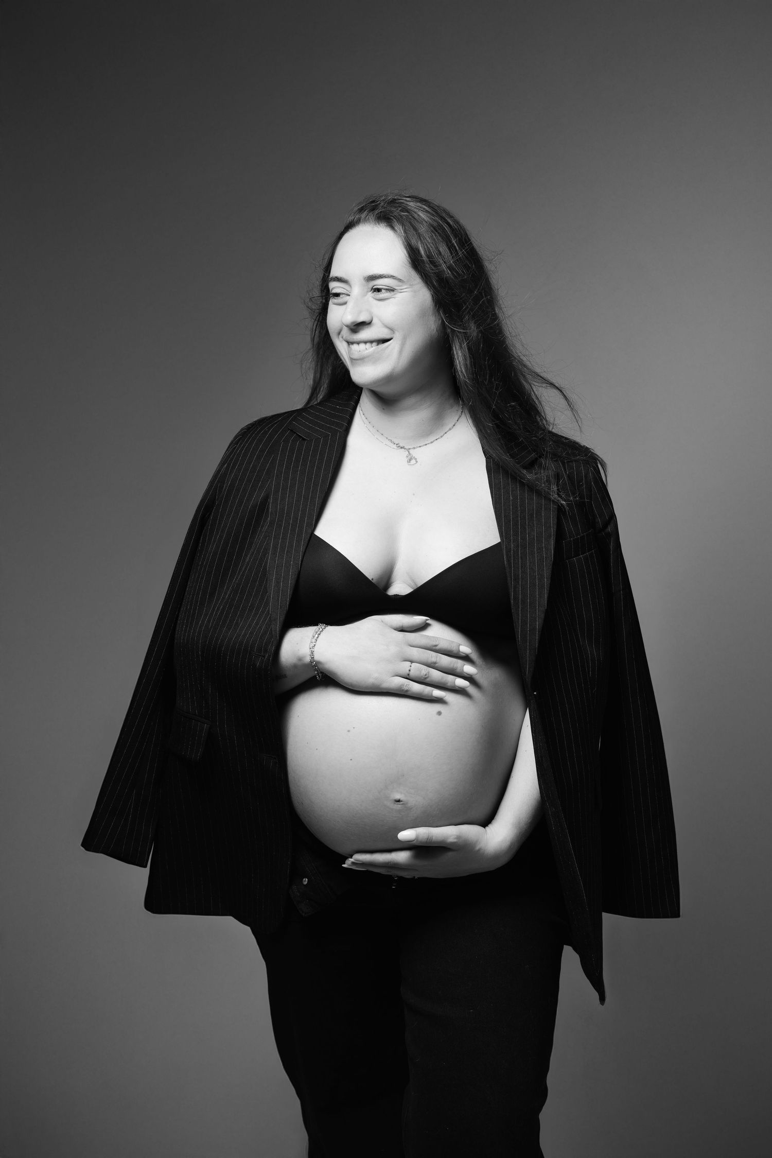 Black and white portrait of a smiling pregnant woman in a blazer cradling her baby bump in a studio.
