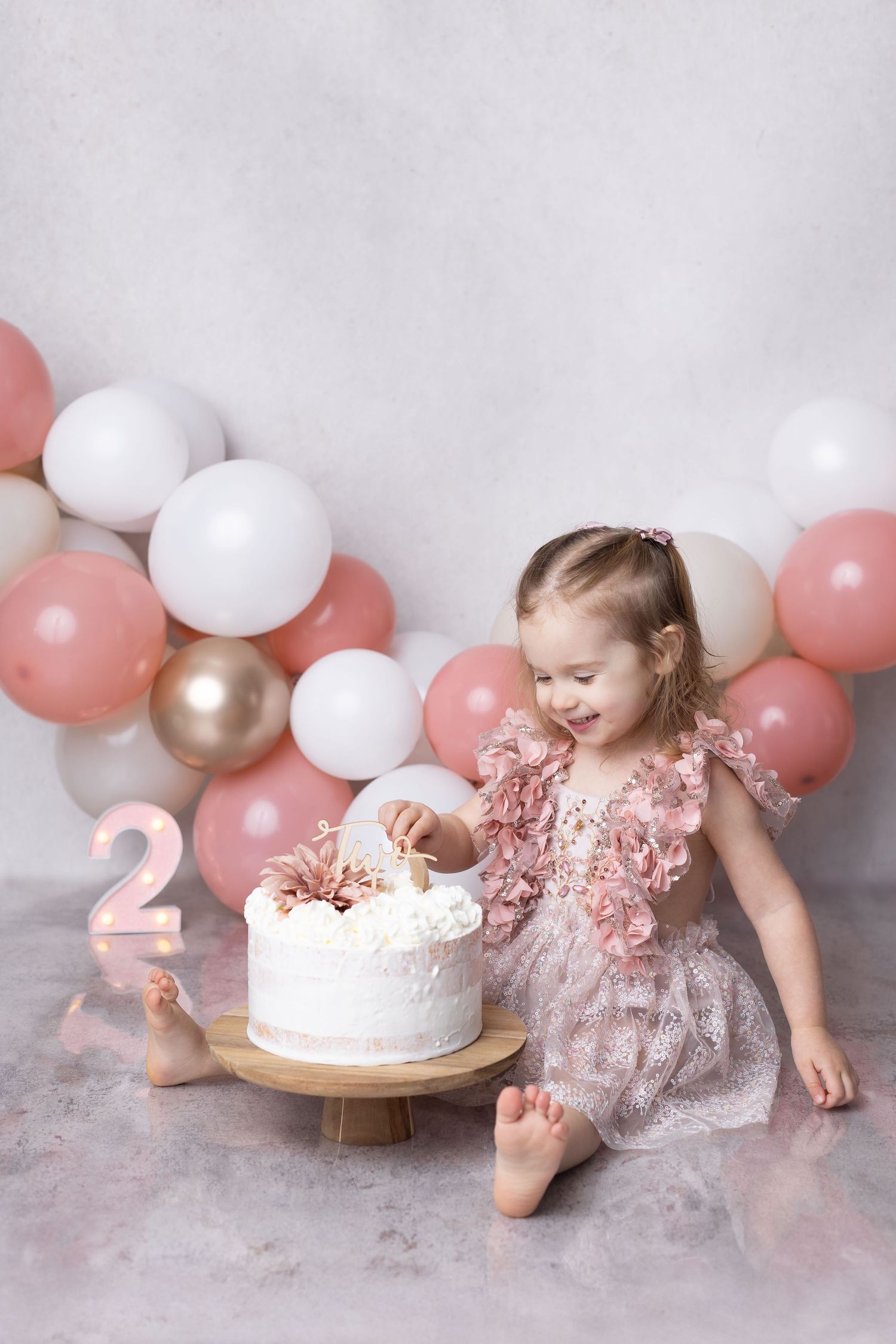 Happy toddler girl in pink floral dress plays with white birthday cake surrounded by pink and white balloons.