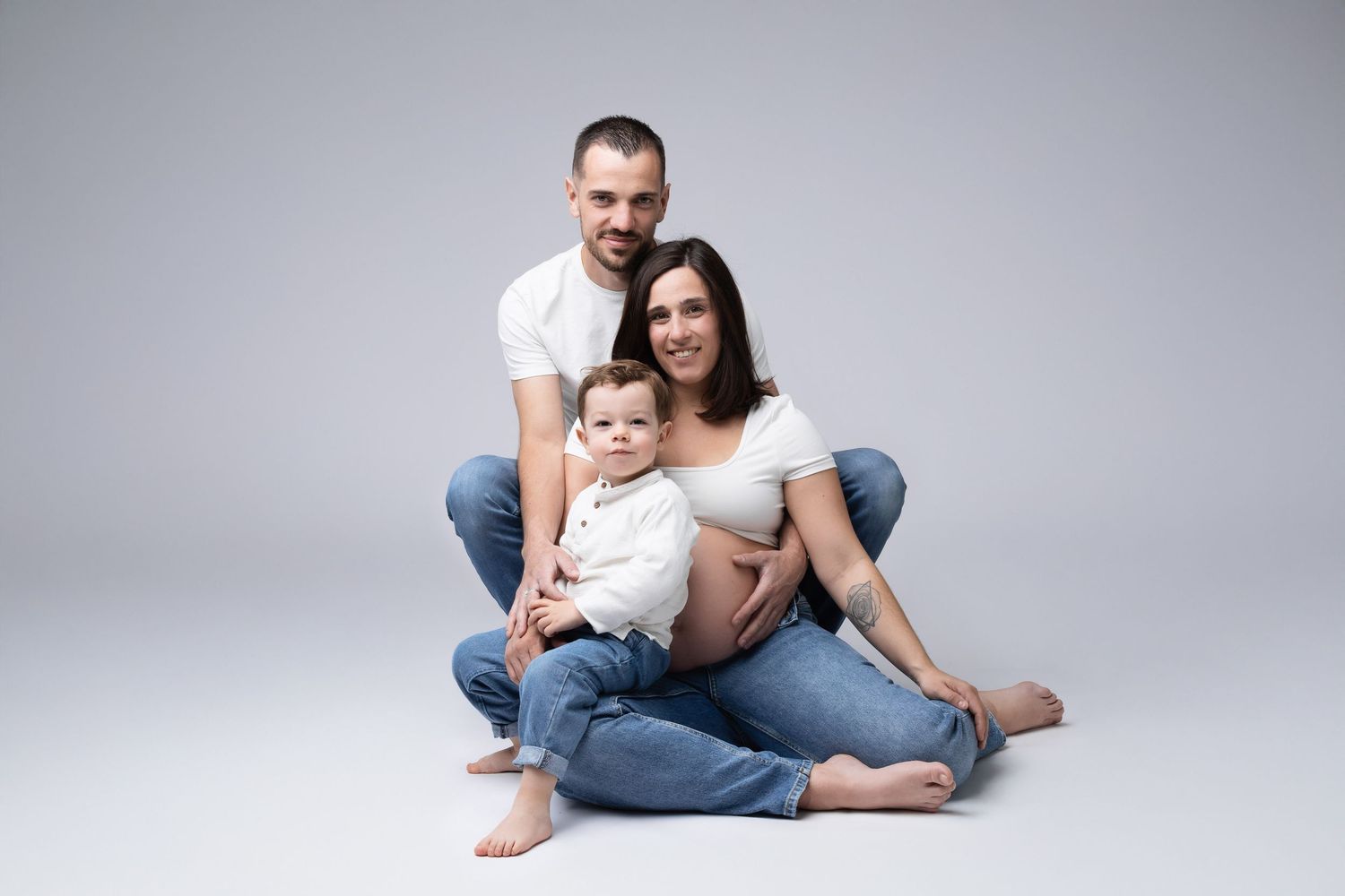 Happy family of three with pregnant mother, father, and baby sitting together on gray background. photographe grossesse chavanoz lyon