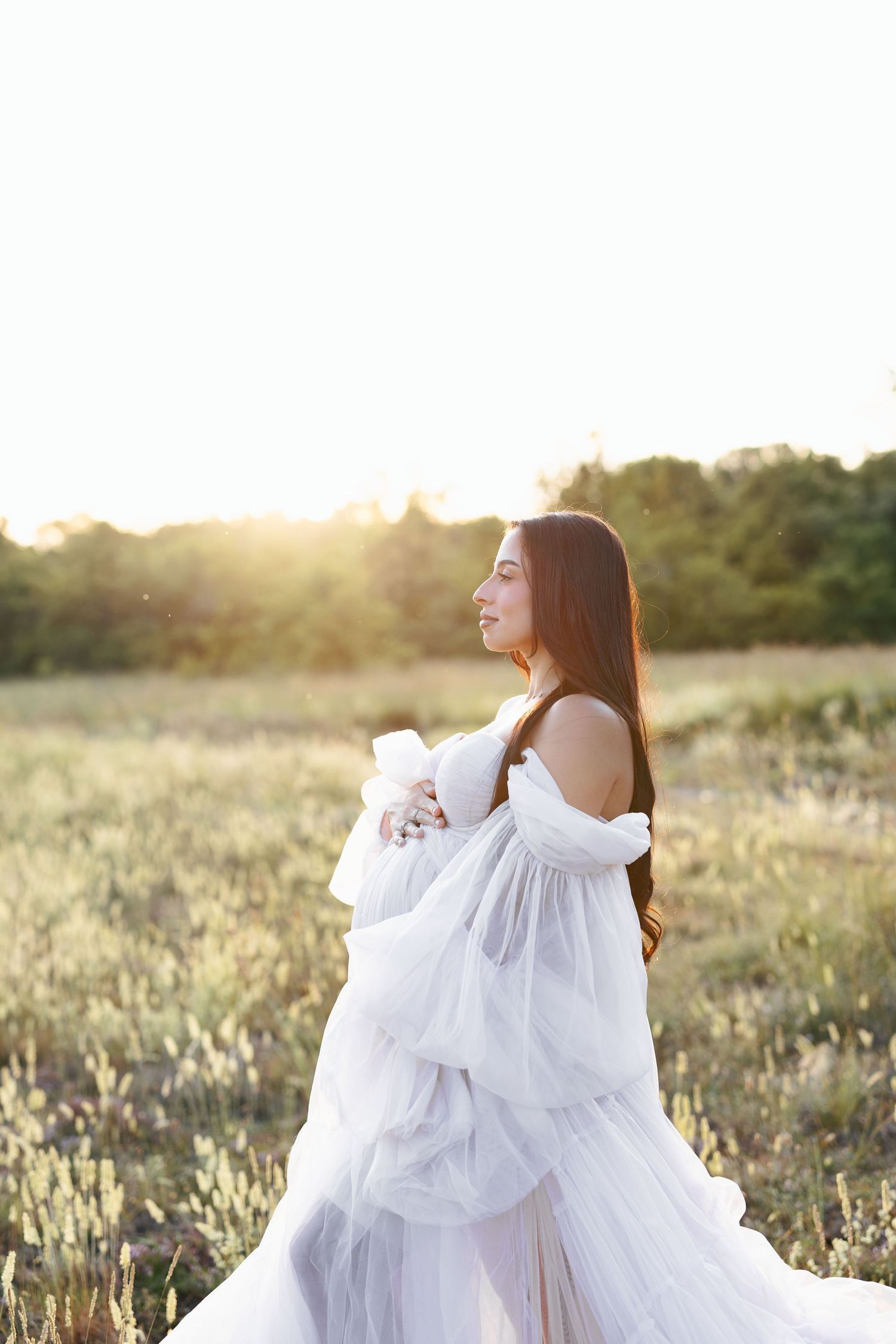 Young woman in white flowing gown standing in sunlit field at golden hour, looking gracefully to the side.