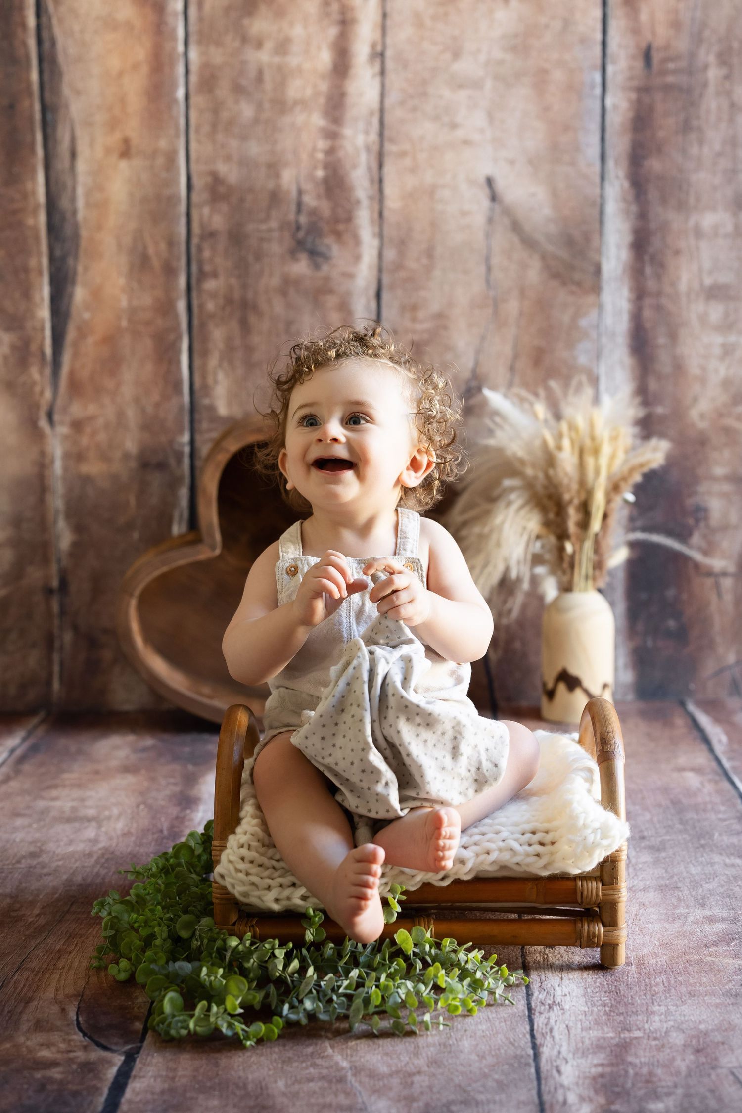 Laughing baby girl in floral romper sitting in wicker basket with greenery on rustic wood floor. photographe grossesse chavanoz lyon