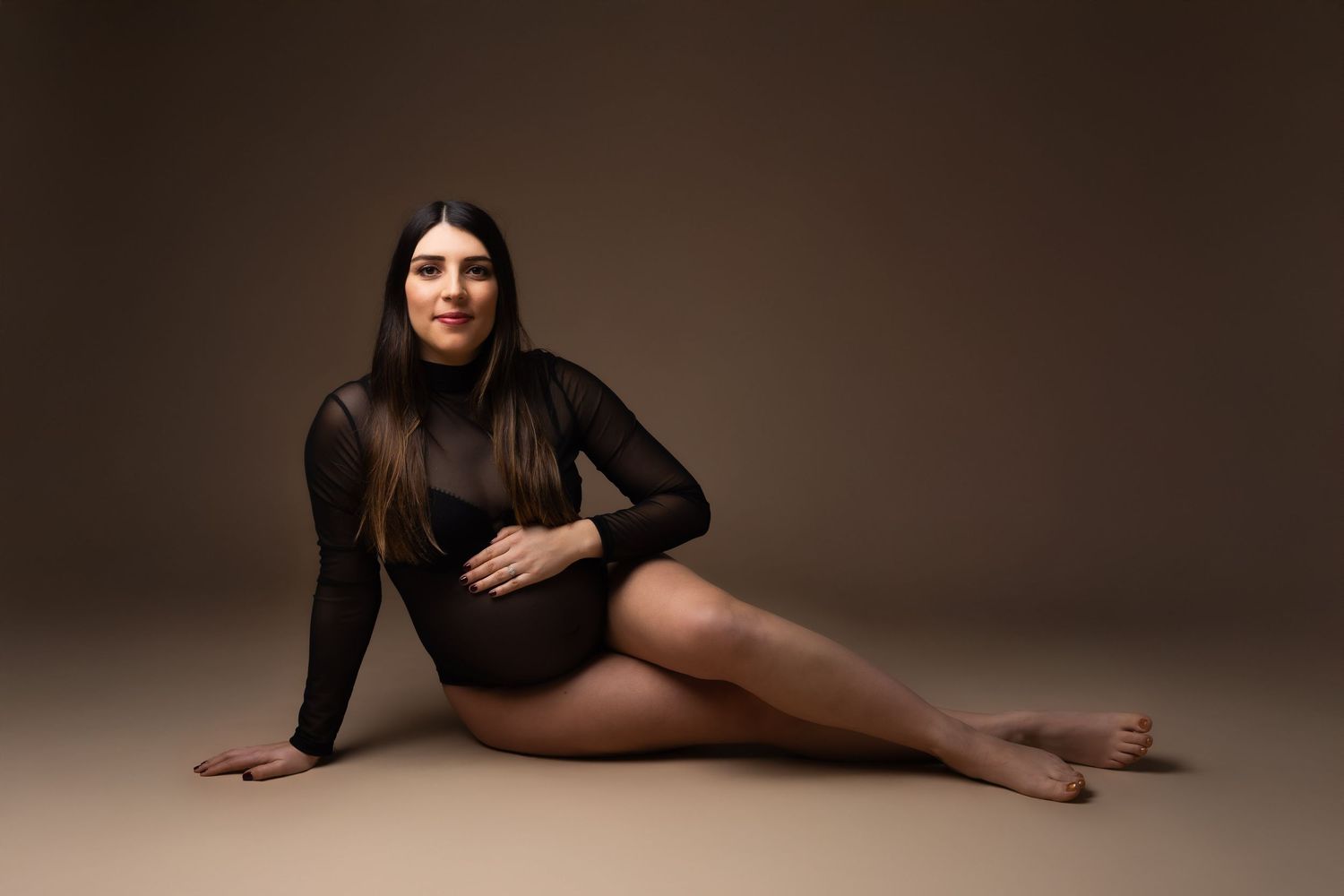 Smiling pregnant woman in black mesh bodysuit sitting gracefully on floor against brown studio background.