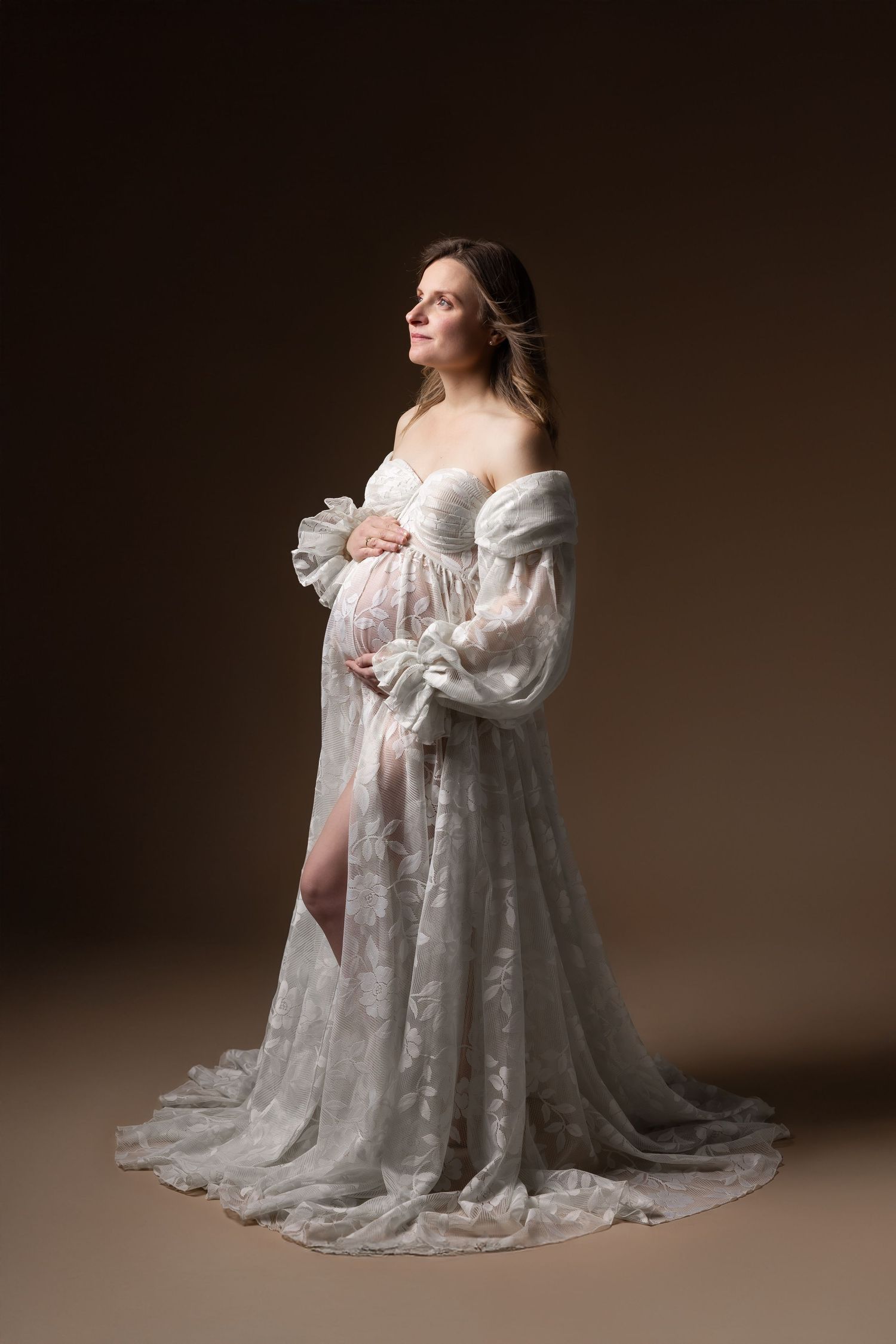 Pregnant woman in elegant white lace off-shoulder gown posing in warm studio setting.