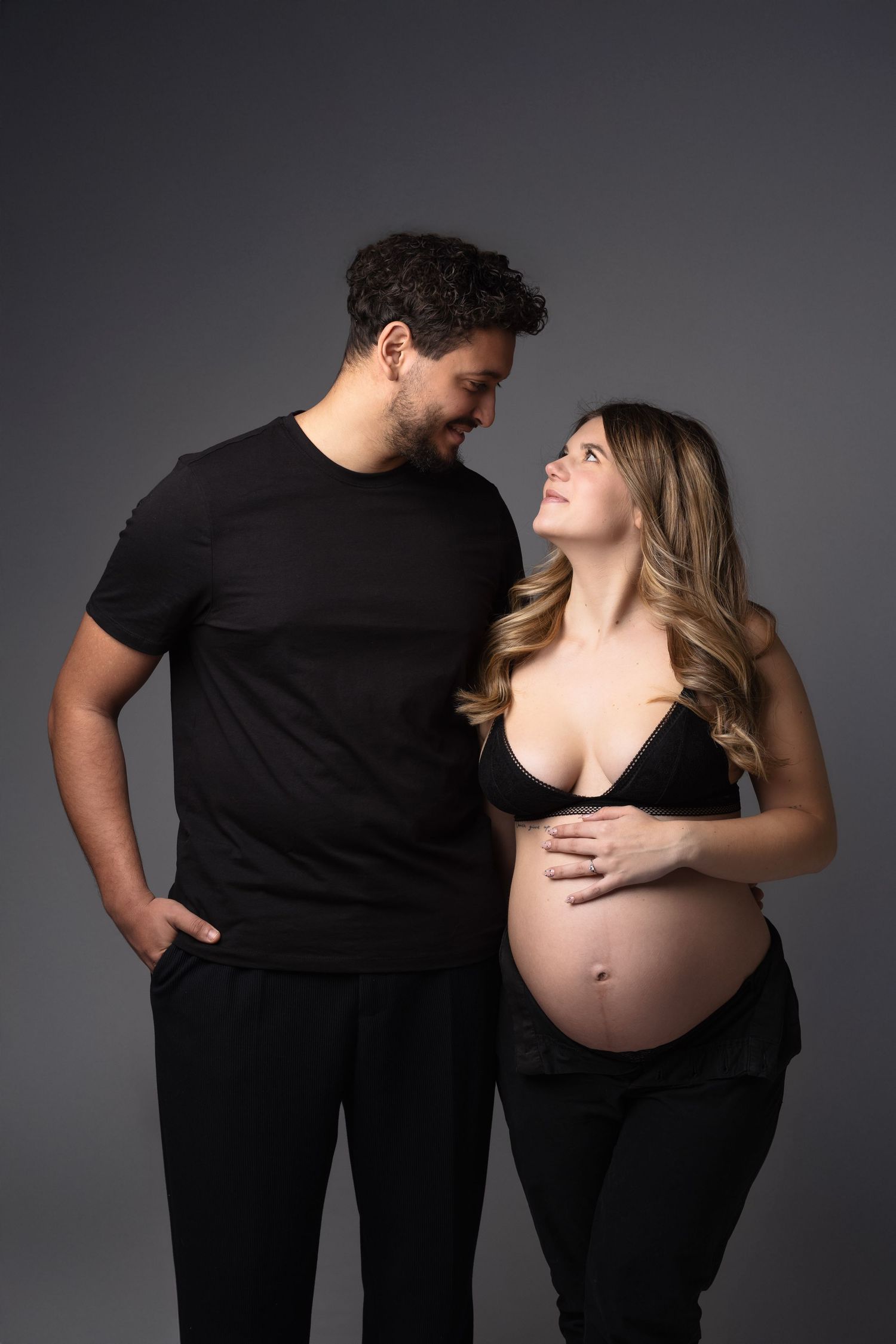 Smiling pregnant couple dressed in black, woman cradling baby bump while looking up at partner in studio. photographe grossesse chavanoz lyo