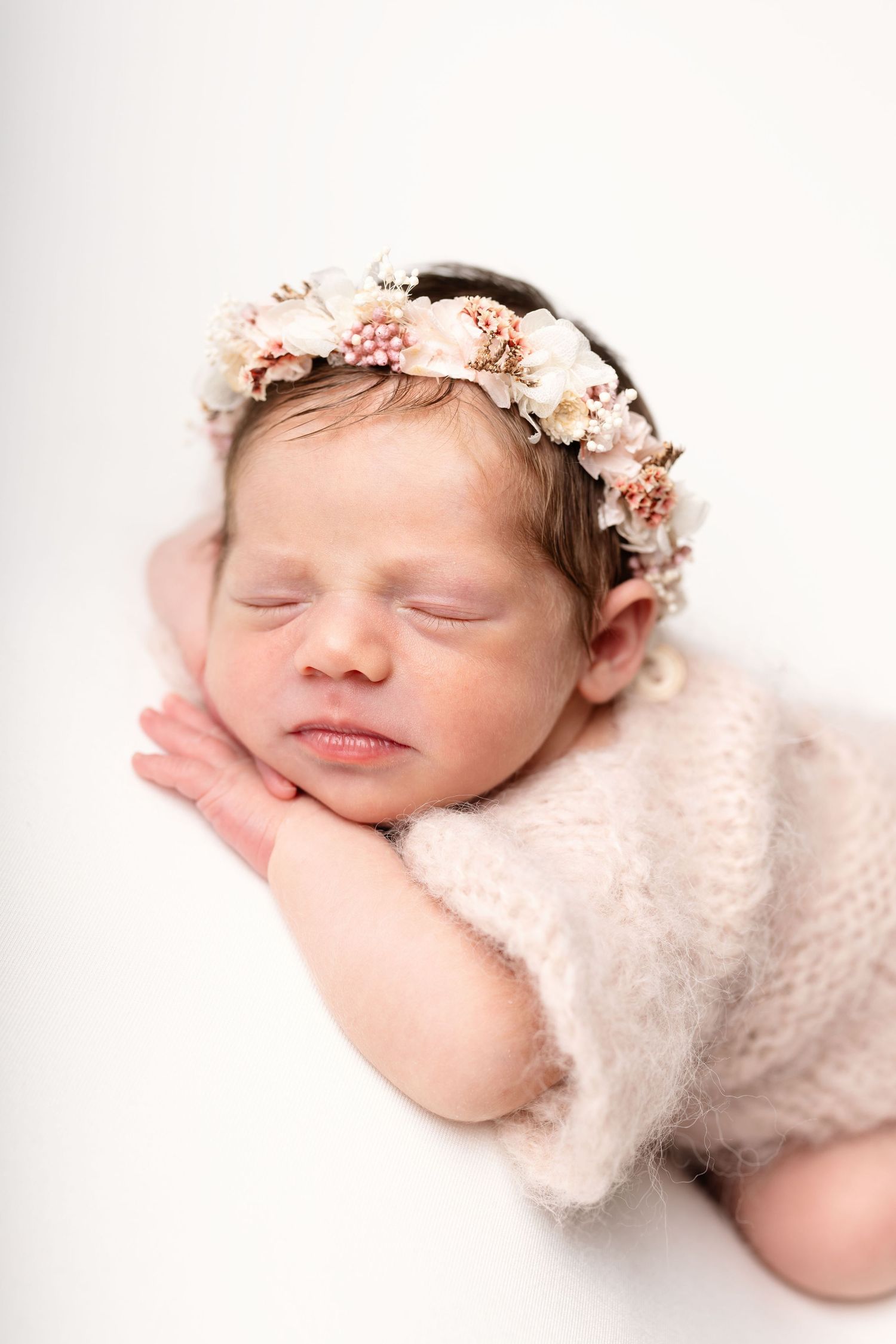 Sleeping newborn baby girl wearing a delicate floral crown and soft pink knit romper on white background.