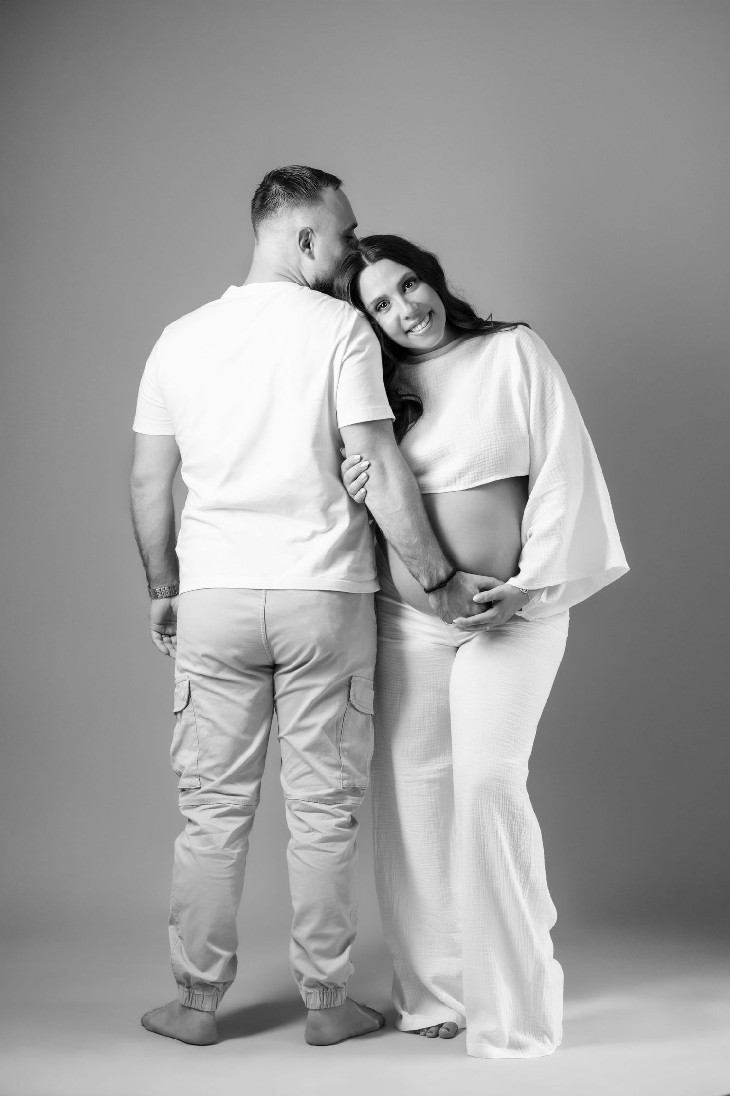 Black and white maternity photo of a couple embracing, woman in white dress leaning on her partners shoulder smiling.