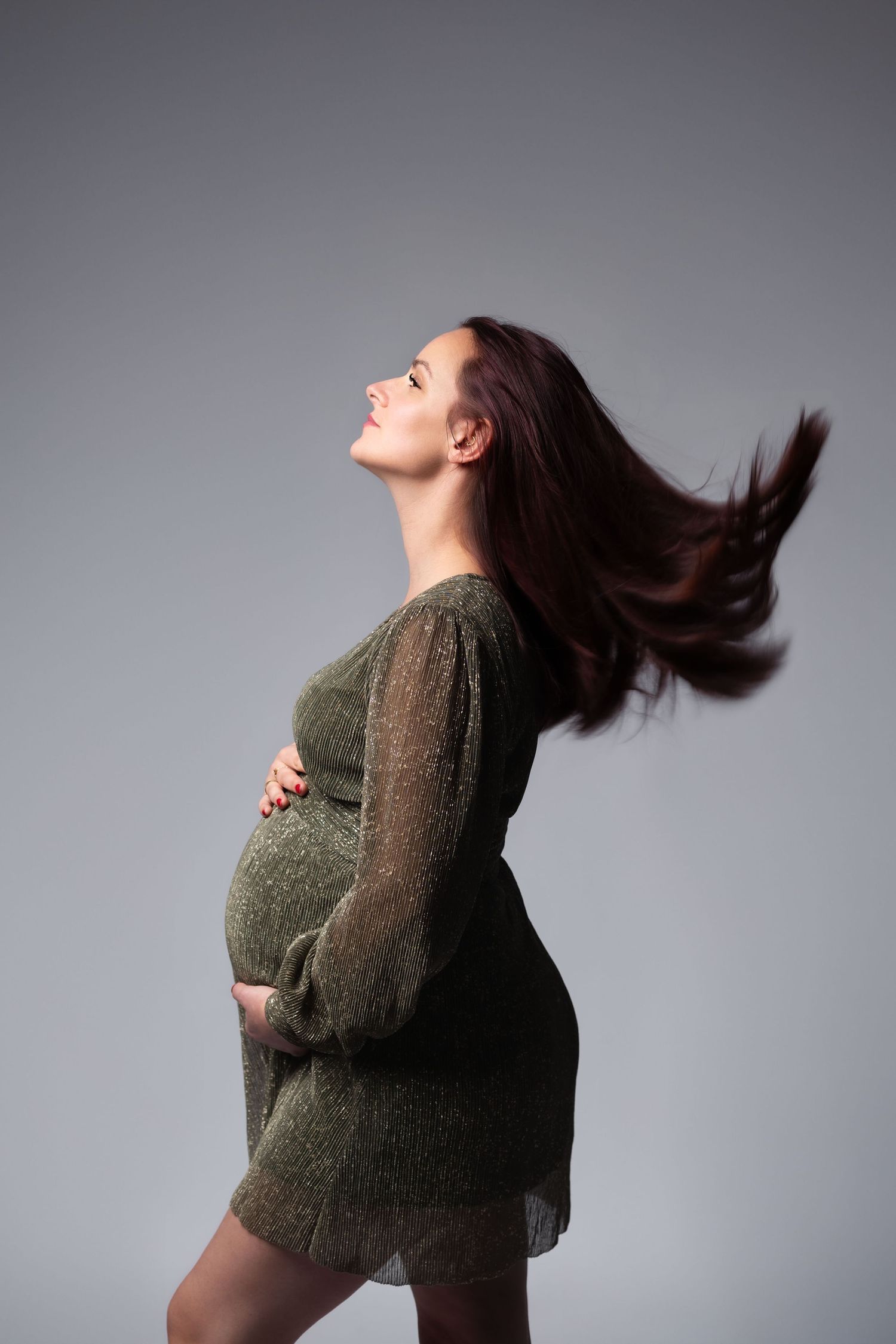 Pregnant woman in a green dress tossing her long hair back, hands on belly in a studio.