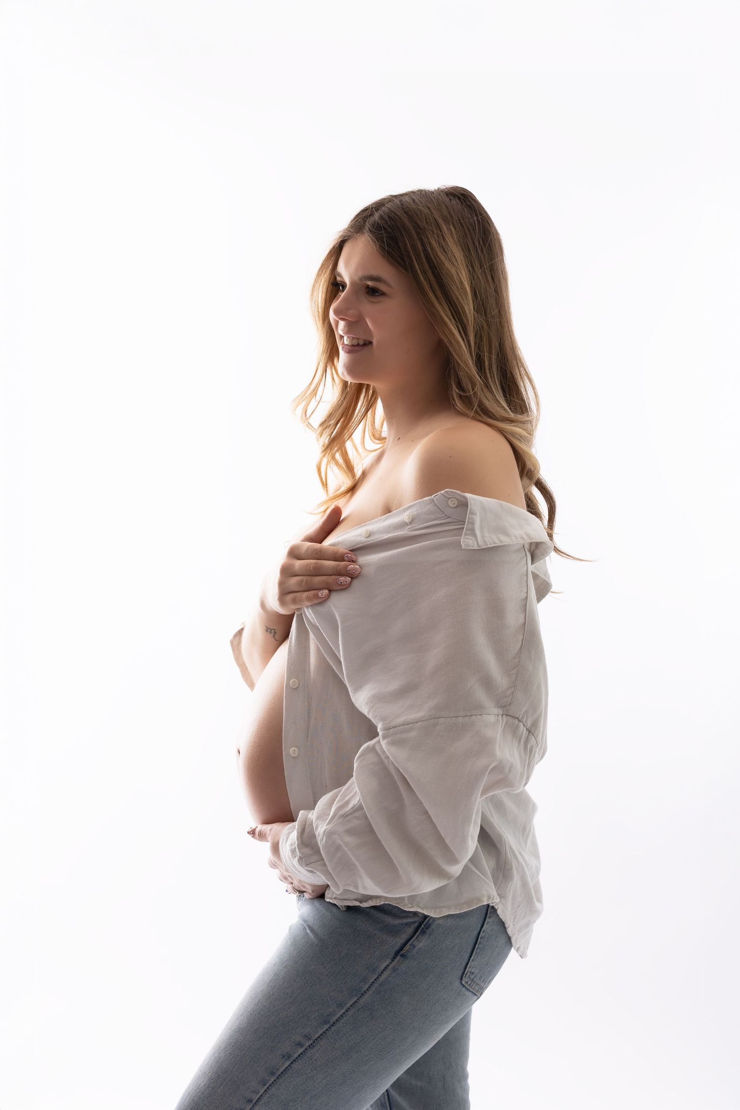 Smiling pregnant woman in open white shirt and jeans revealing her baby bump in studio. photographe grossesse chavanoz lyon