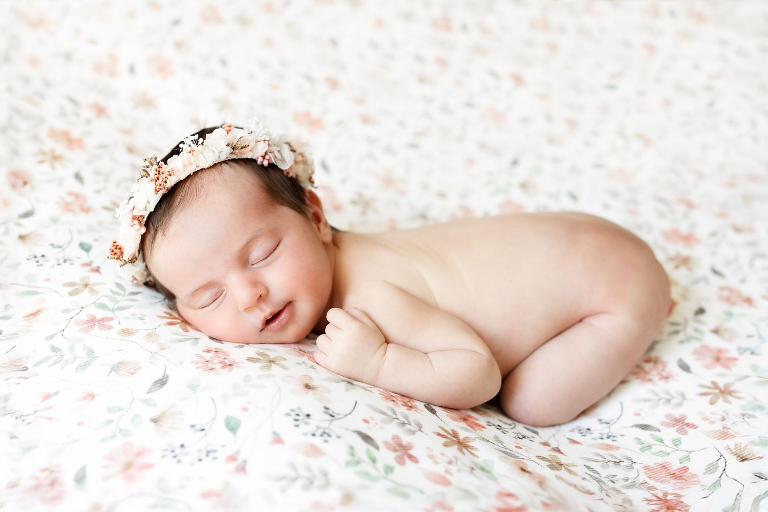 Peaceful newborn baby girl sleeping curled up on floral blanket wearing a delicate flower headband.