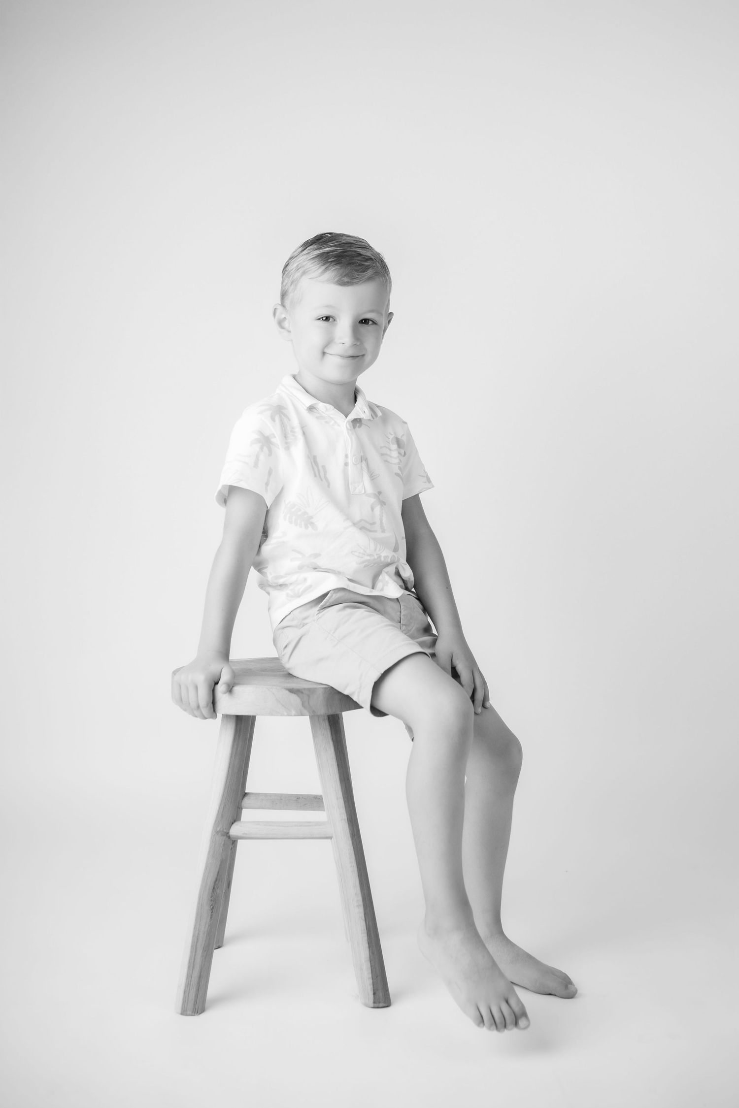A smiling young boy sits barefoot on a wooden stool in a black and white studio portrait.
