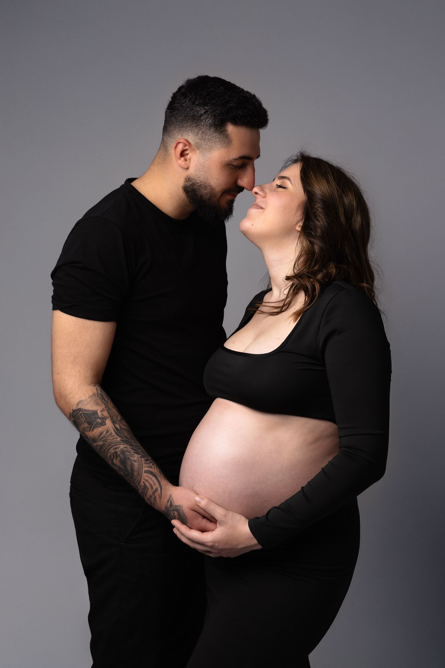 Couple in matching black outfits sharing tender moment while cradling pregnant belly on grey background. photographe grossesse chavanoz lyon