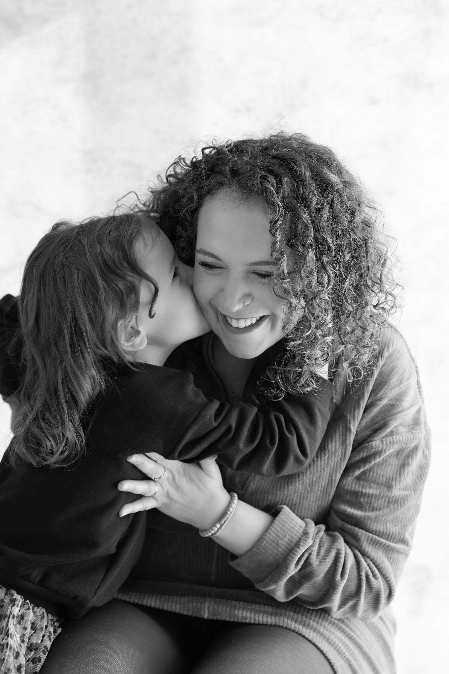 Black and white photo of a young child kissing a laughing curly-haired mother on the cheek.
