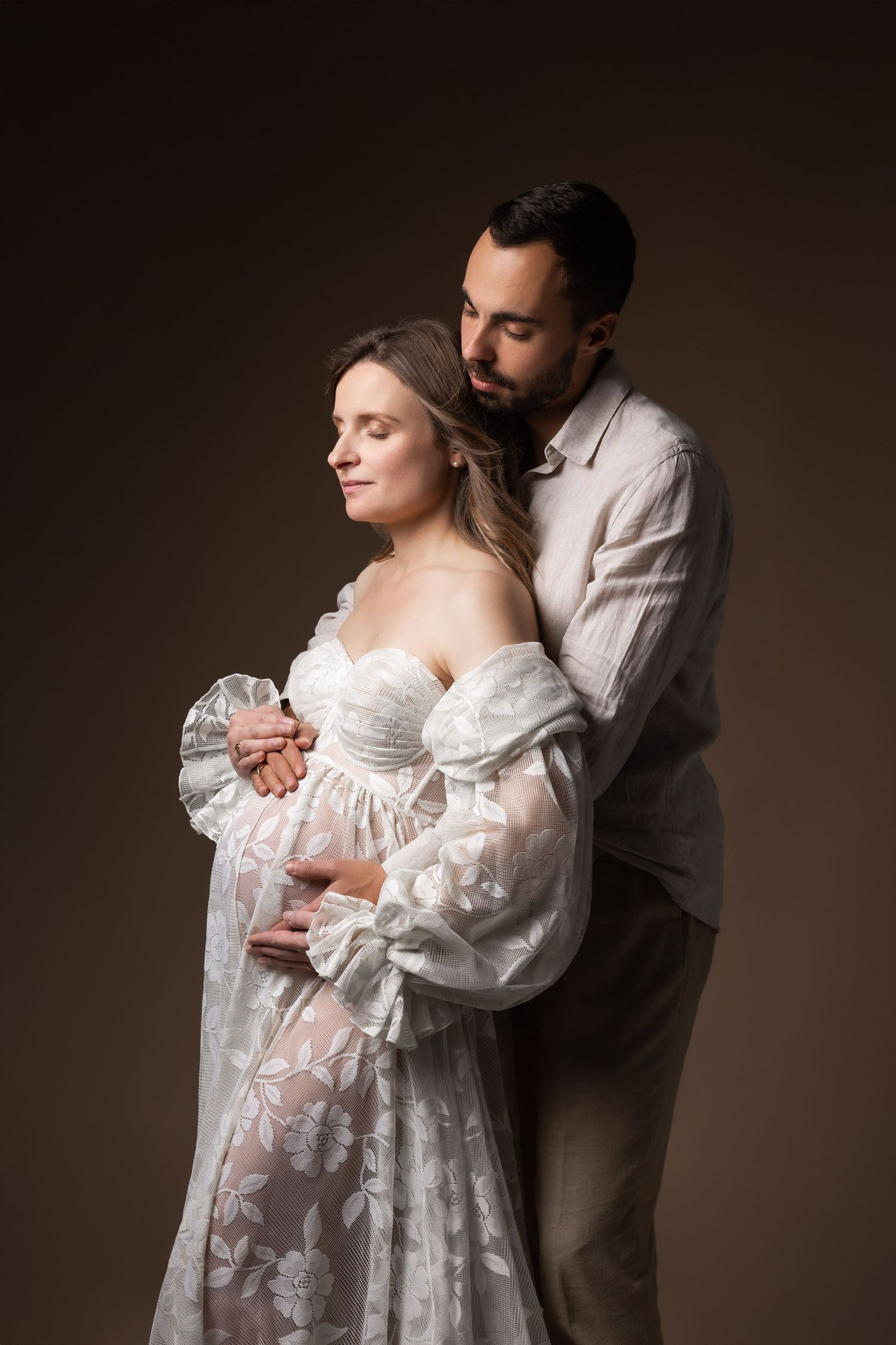 Man tenderly embracing pregnant woman in white lace gown in romantic studio portrait.