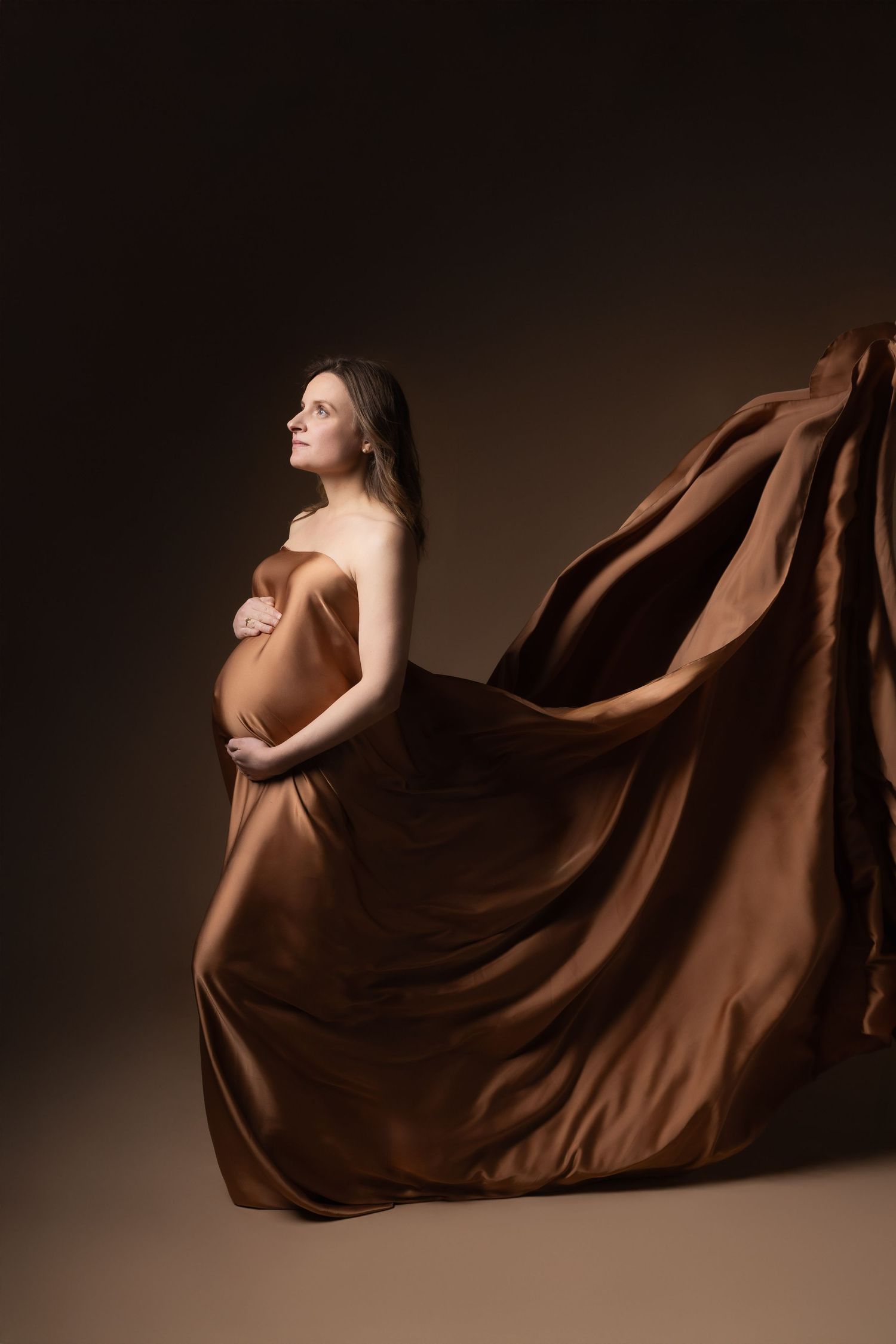 Pregnant woman wrapped in flowing bronze silk fabric posing in dramatic studio portrait.
