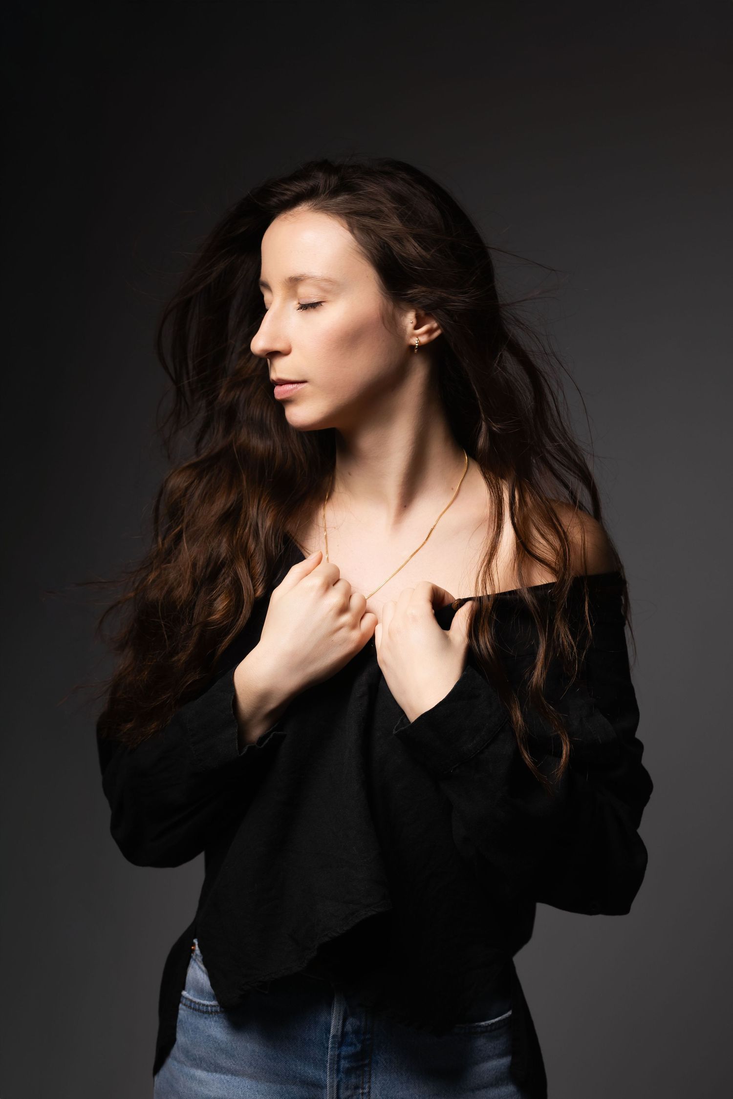 Young woman with long wavy brunette hair in black top holds necklace with eyes closed on dark background.