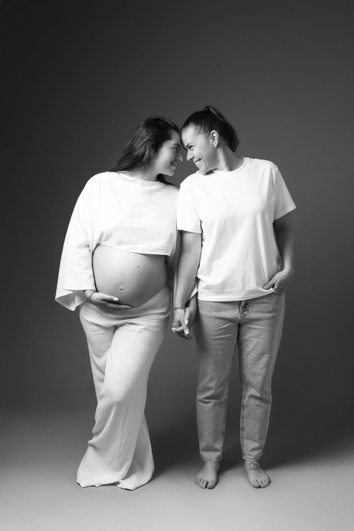 Black and white photo of two women holding hands, one visibly pregnant, wearing white outfits in a studio setting.
