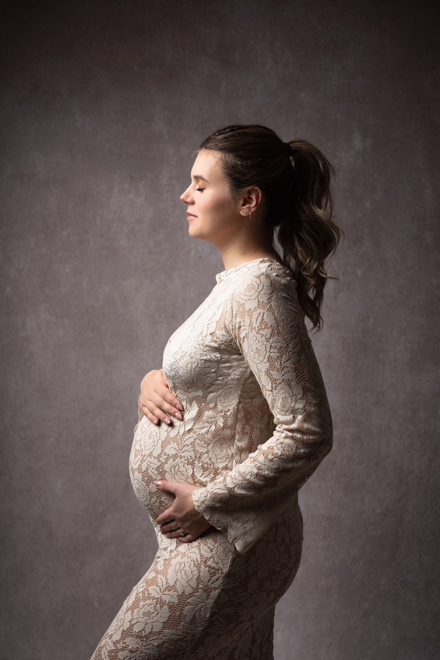 Pregnant woman in white lace dress holding baby bump, eyes closed, side profile against gray textured background.