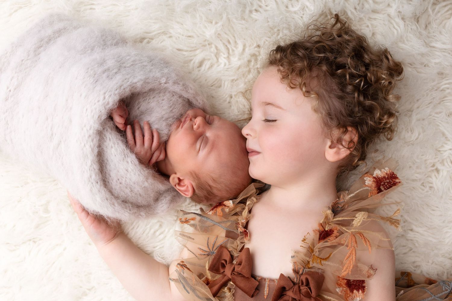 Adorable curly-haired toddler girl gently kissing sleeping newborn sibling wrapped in gray knit on fur blanket. photographe naissance