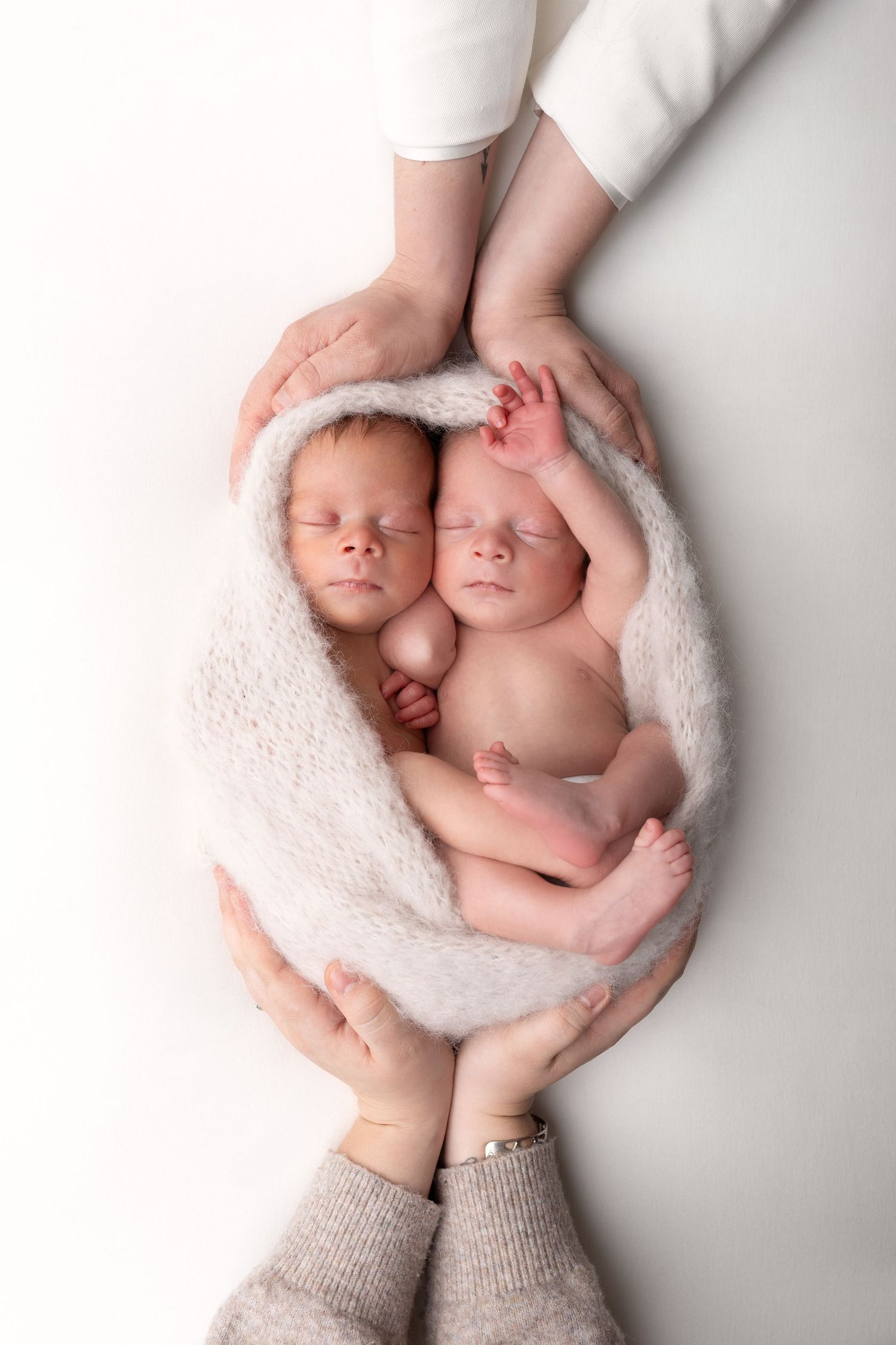 Two sleeping newborn twins wrapped in a soft white blanket, cradled gently by parents hands on a white background.
