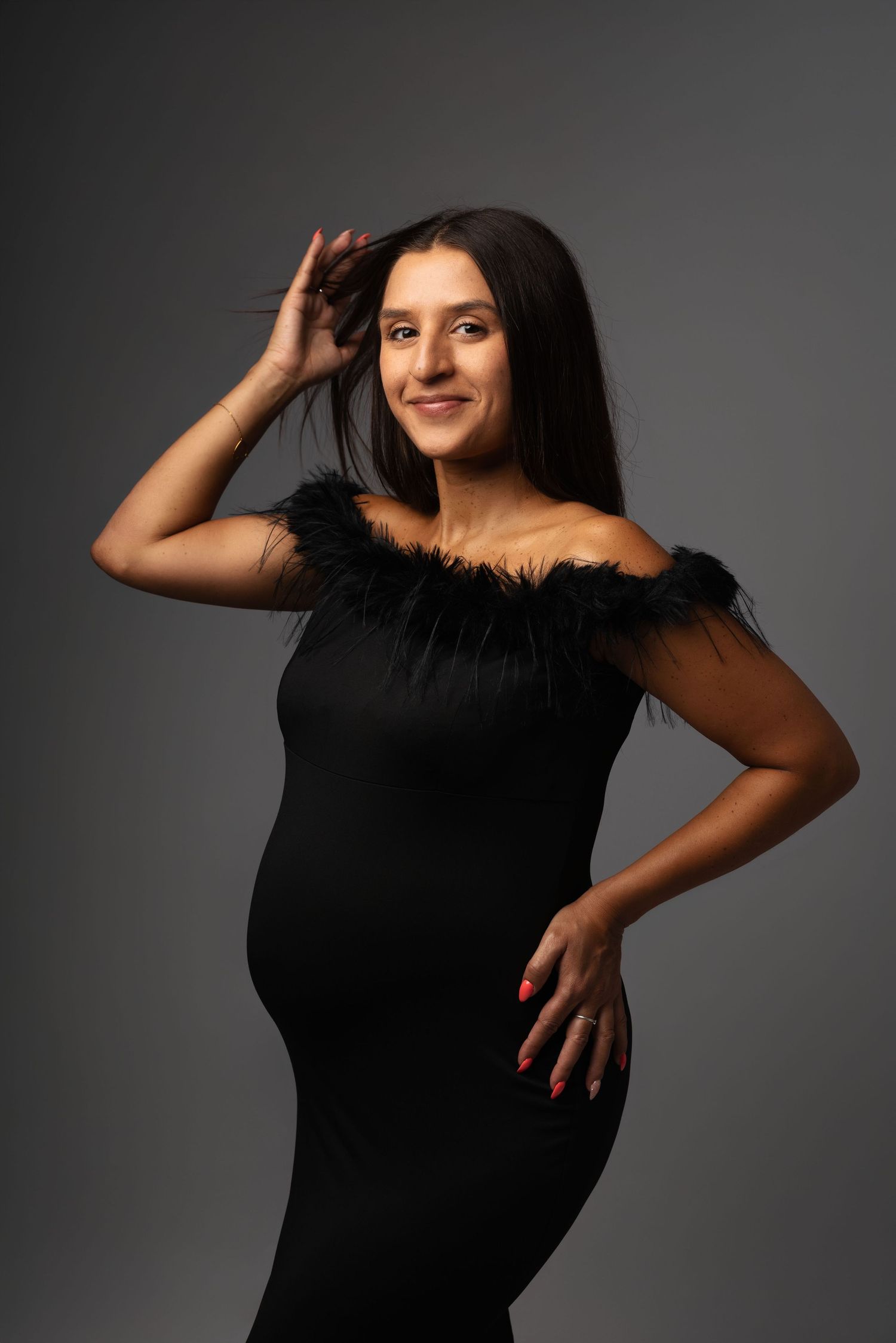 Pregnant woman in elegant black off-shoulder dress with feather trim, posing gracefully against gray background.