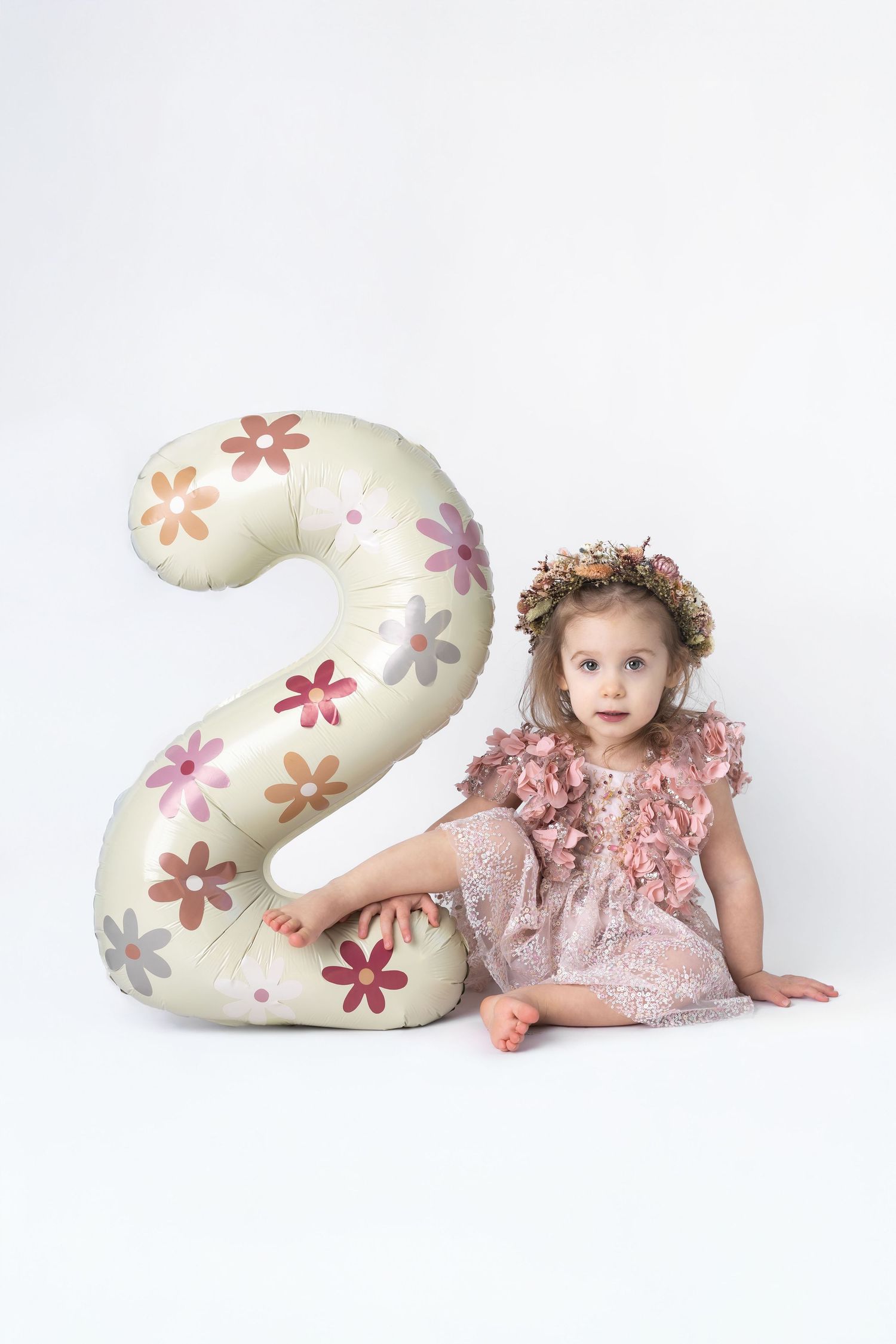Cute toddler girl in pink dress and floral crown sits beside a large decorative number 2 balloon.
