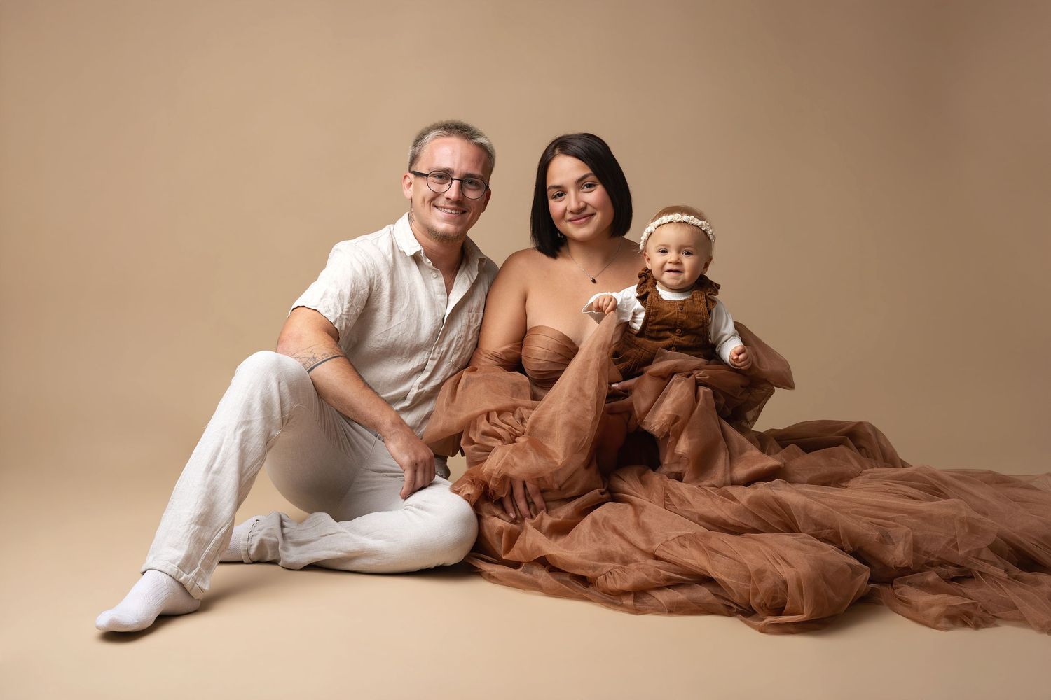 Family of three posing for a studio maternity photo, parents smiling with toddler on beige background with brown fabric.